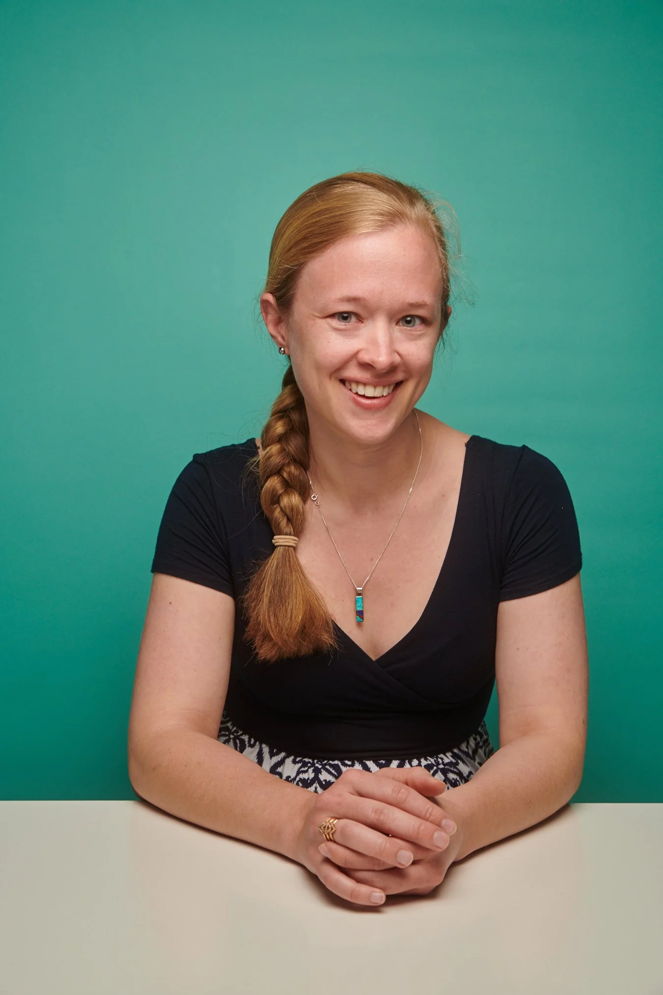A smiling woman with red hair in a braid, wearing a black top, a necklace with a rectangular pendant, and earrings. She is sitting at a white table with her hands folded, against a teal background.
