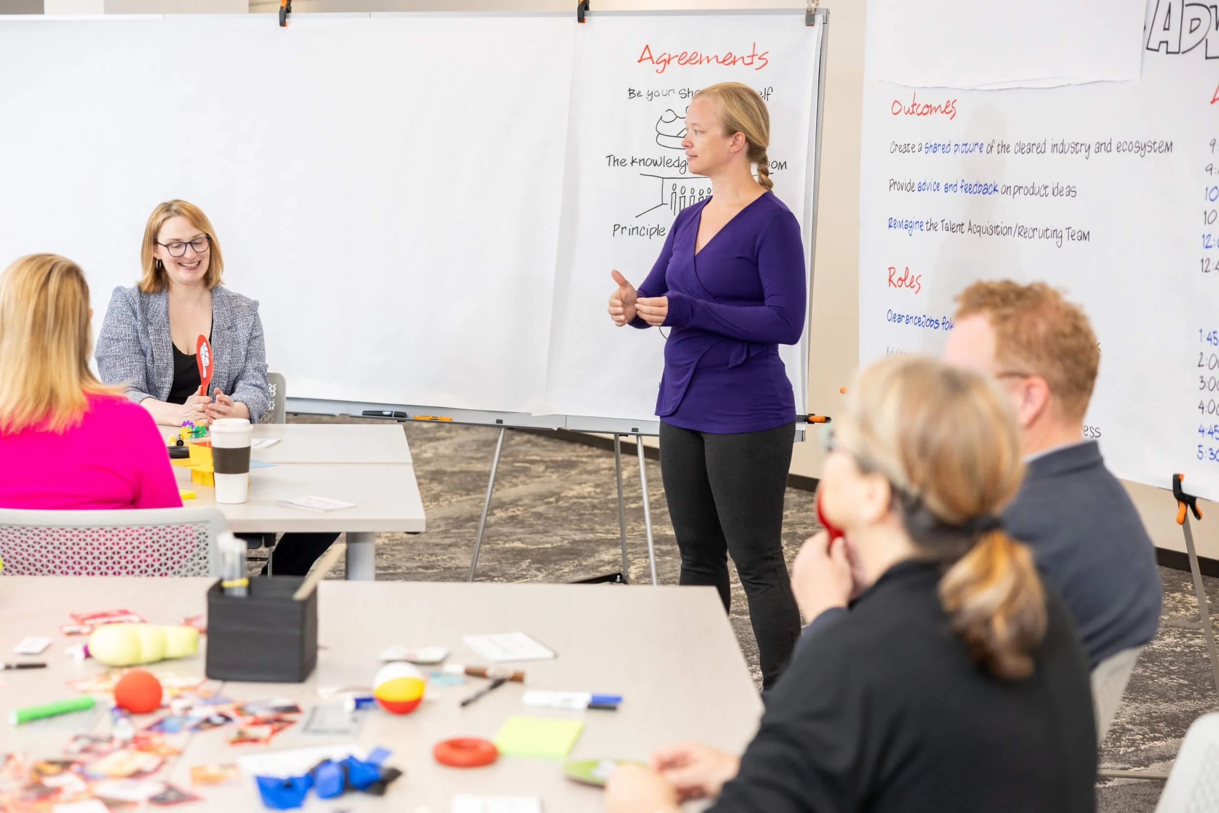 A woman in a purple shirt facilitating to a group of people in a classroom or workshop setting, with whiteboards and notes behind her.