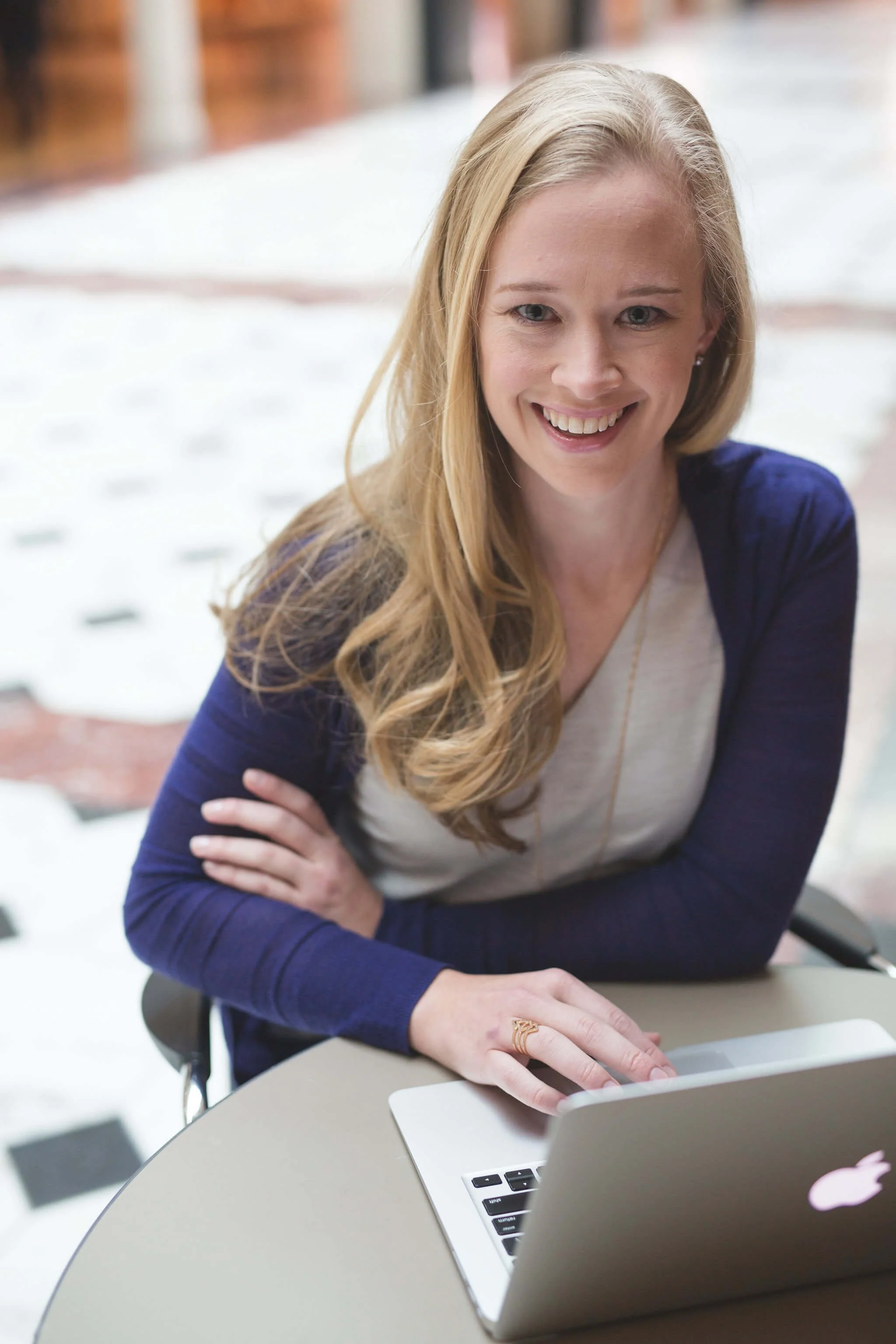 A woman with blonde hair smiles while sitting at a table using a laptop in a bright indoor setting.