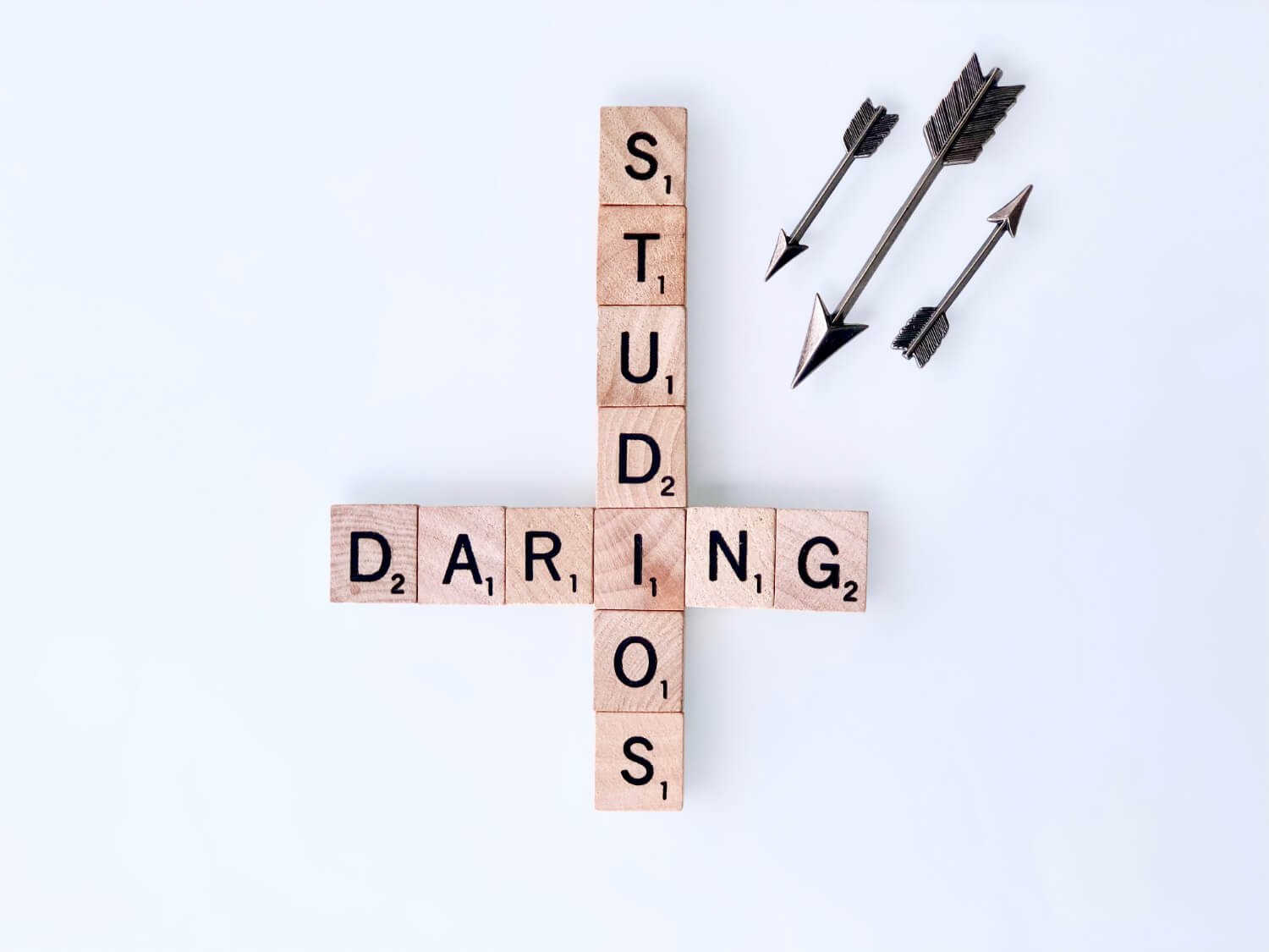 Wooden Scrabble tiles arranged to spell the words 'STUDIOS' vertically and 'DARING' horizontally, forming a cross, with three metal arrows lying on a white surface.
