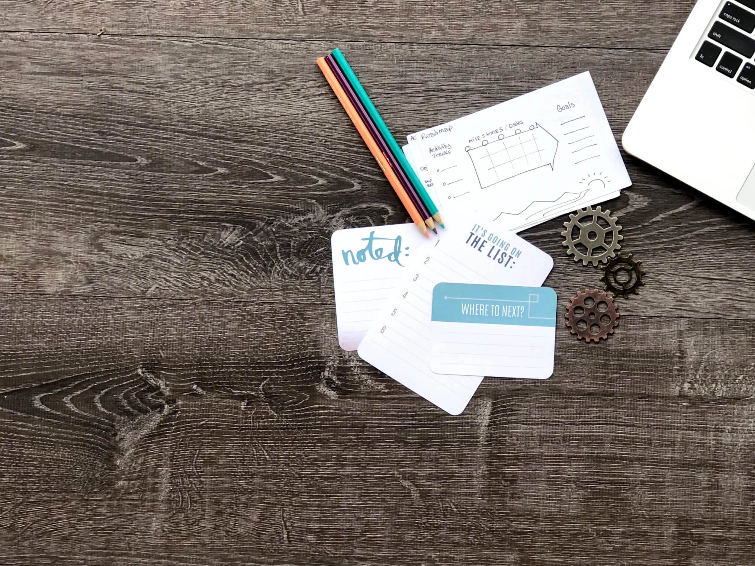 A wooden desk with a laptop, a stack of papers with handwritten notes and a small doodle, colored pencils, and some metal gears.