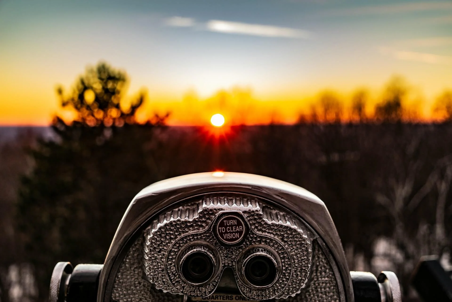 Close-up of a coin-operated binocular viewer facing a sunset with a blurred background of trees and sky.