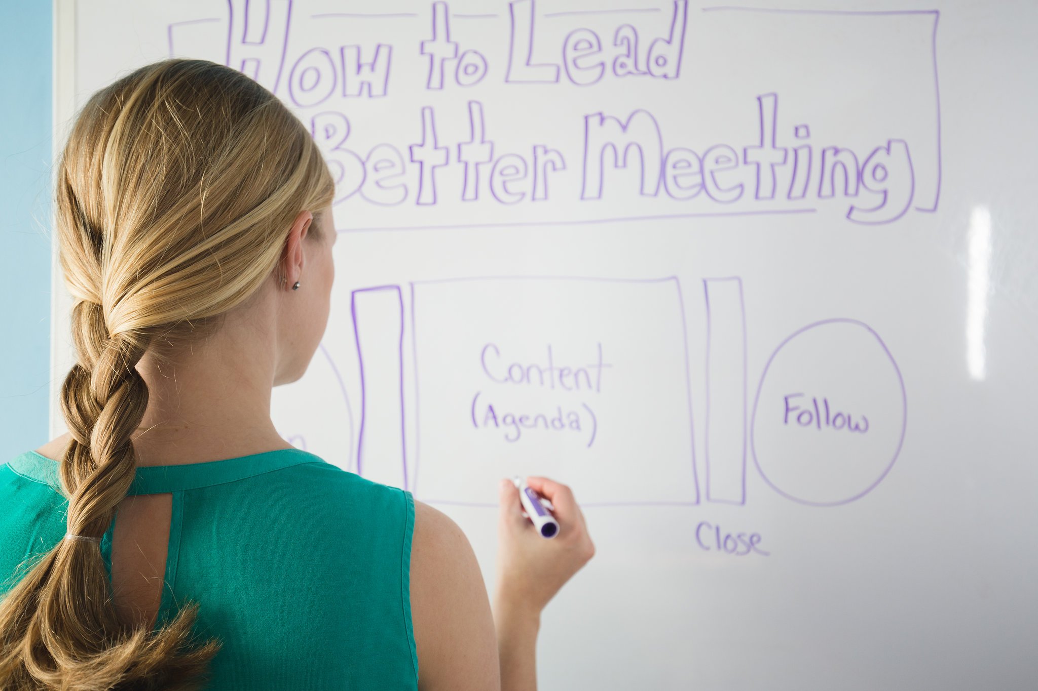 A woman with long wavy blonde hair in a braid, wearing a green top, writing on a whiteboard with a purple marker.
