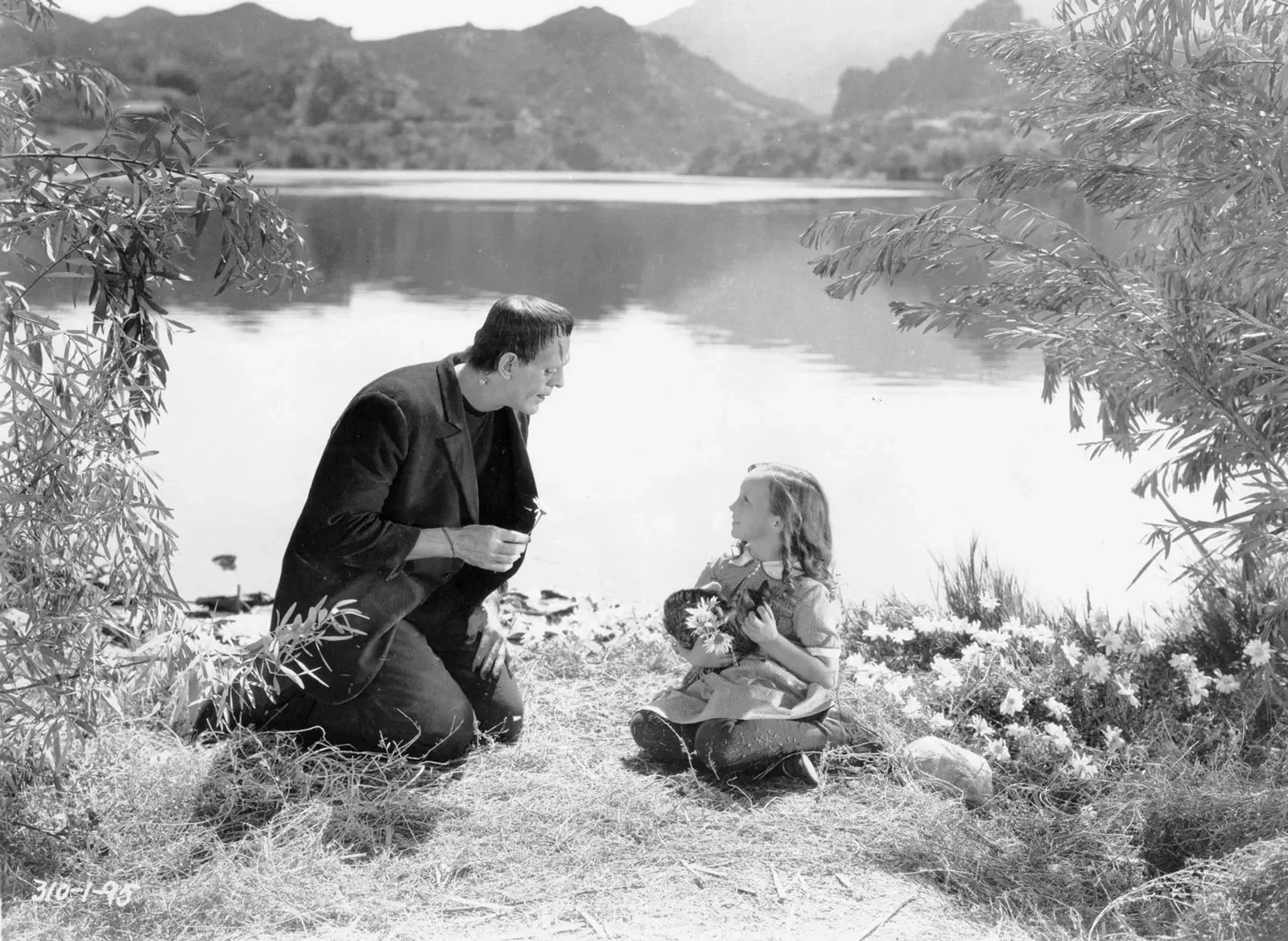 Frankenstein holding a flower kneels in front of a lake with a young girl