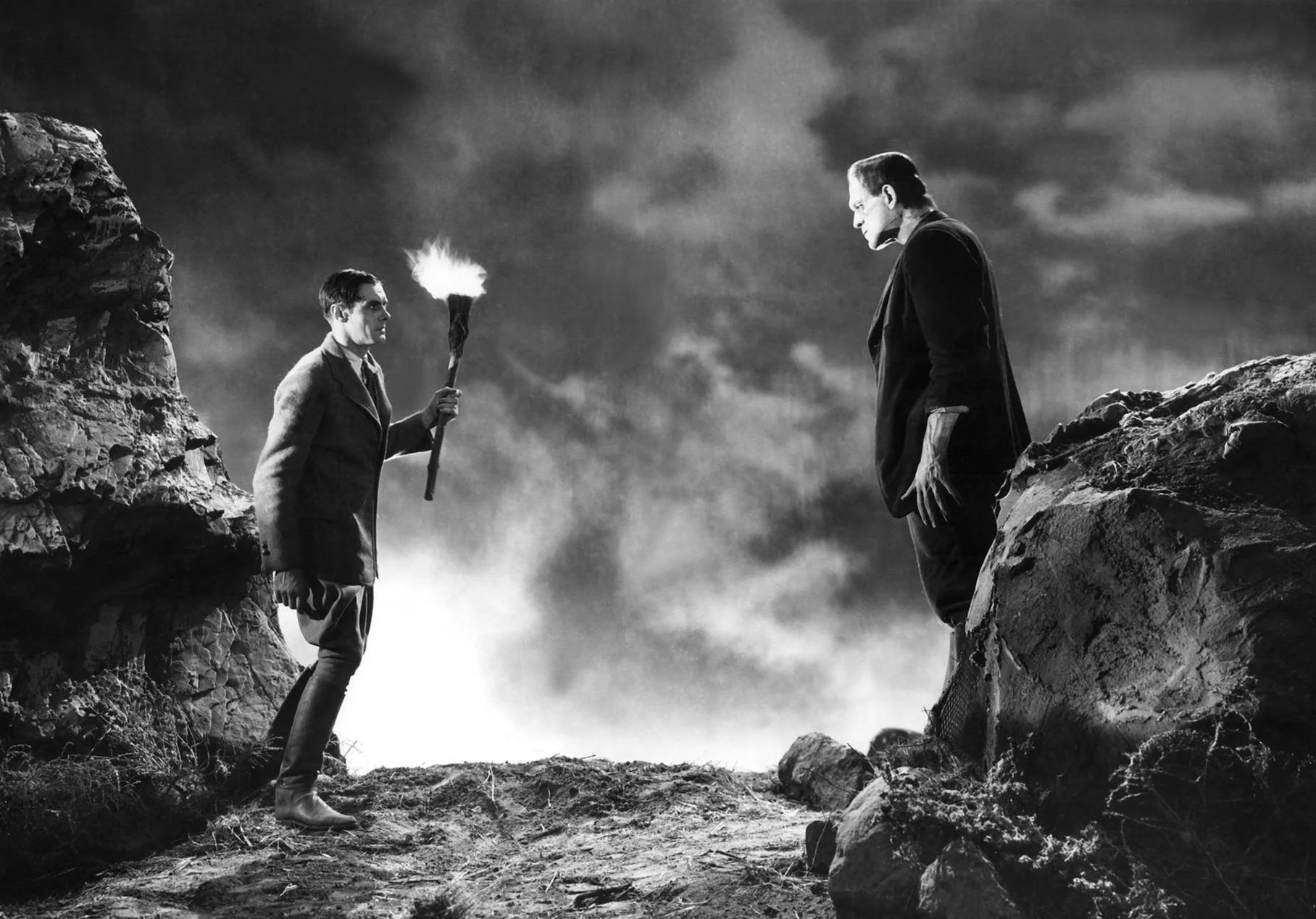 In a rocky terrain, Dr. Frankenstein holds a torch while standing opposite his monstrous creation