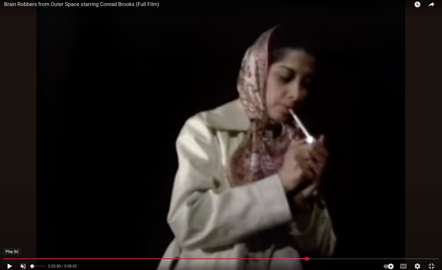 A screenshot gtom Brain Robbers from Outer Space that shows the author of the piece in a head scarf and overcoat lighting a cigarette against a black background