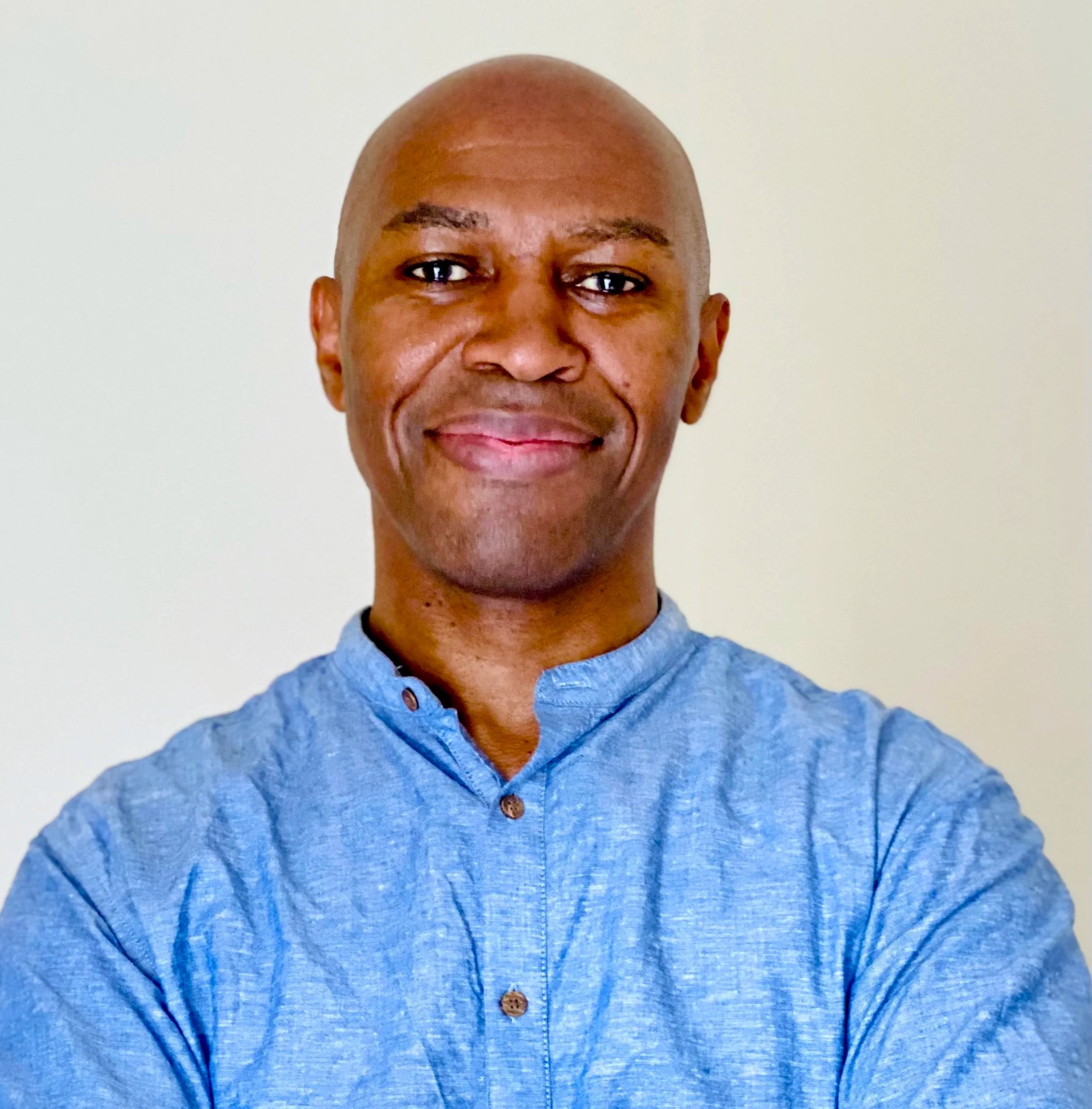 Close-up of a smiling man with a shaved head wearing a blue button-up shirt against a plain white background.