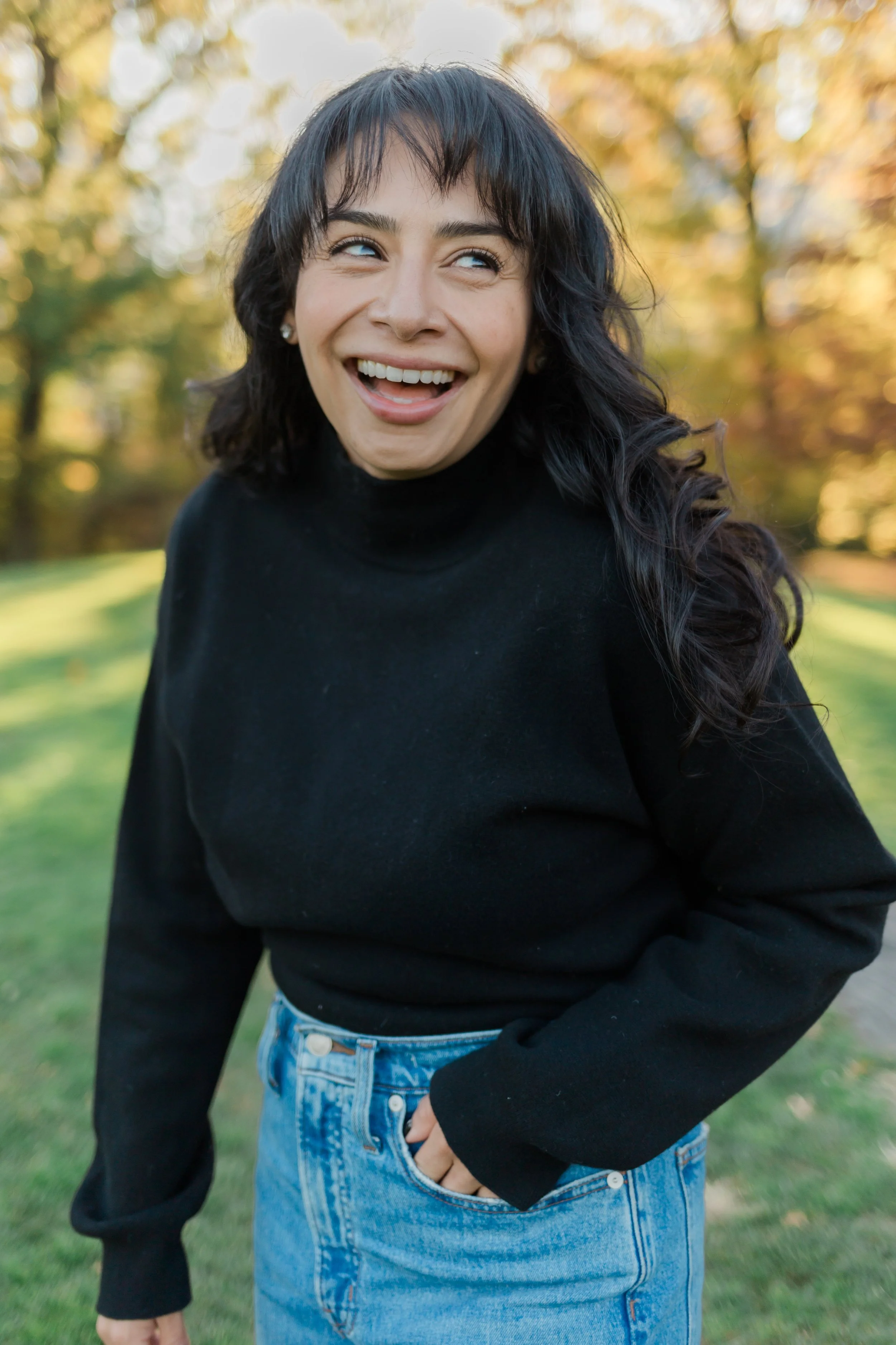 Woman smiling outdoors in autumn, wearing a black turtleneck and blue jeans with trees in the background.
