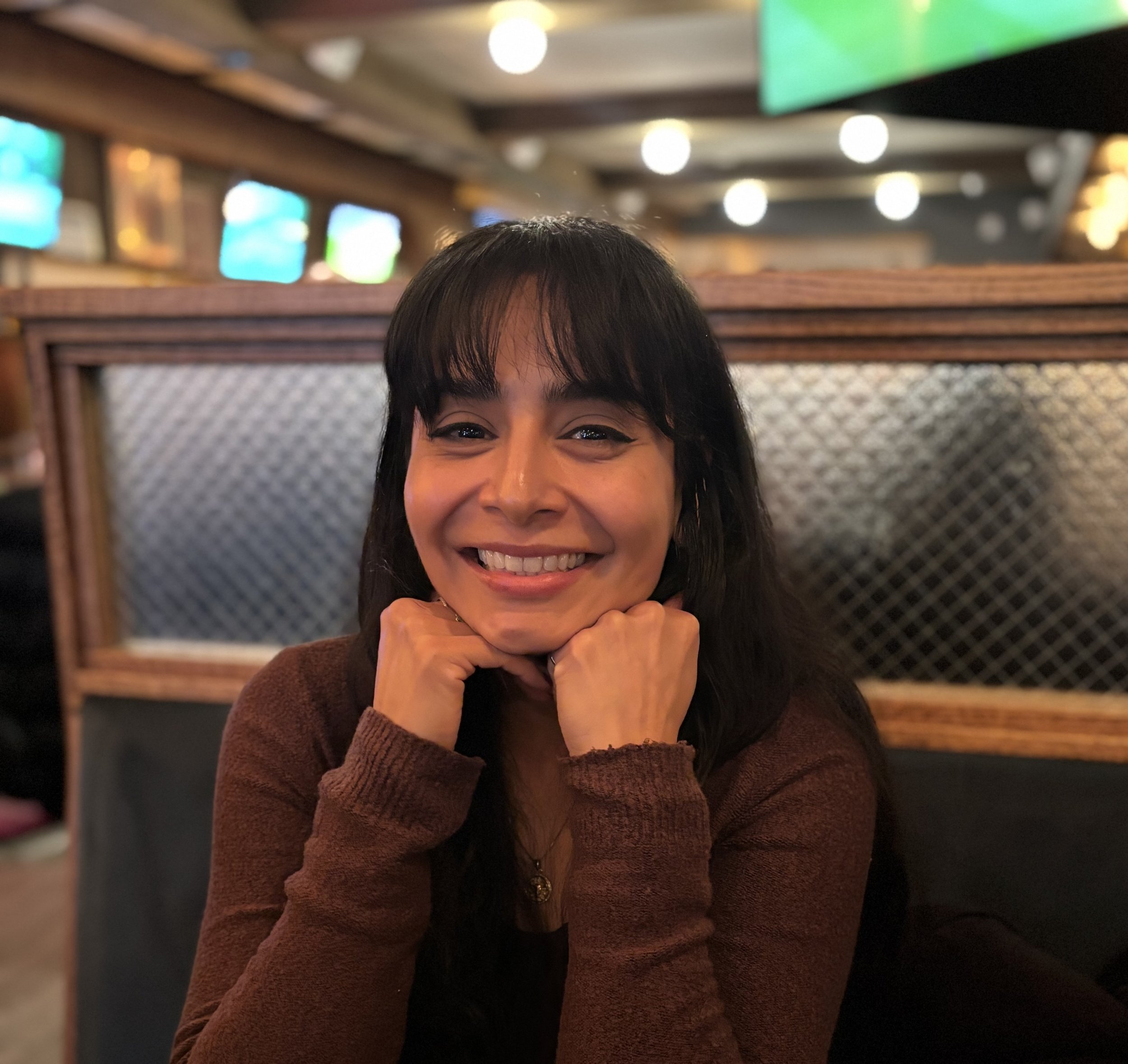 A smiling woman with dark hair and bangs, resting her chin on her hands, seated in a restaurant with dim lighting and wooden decor.