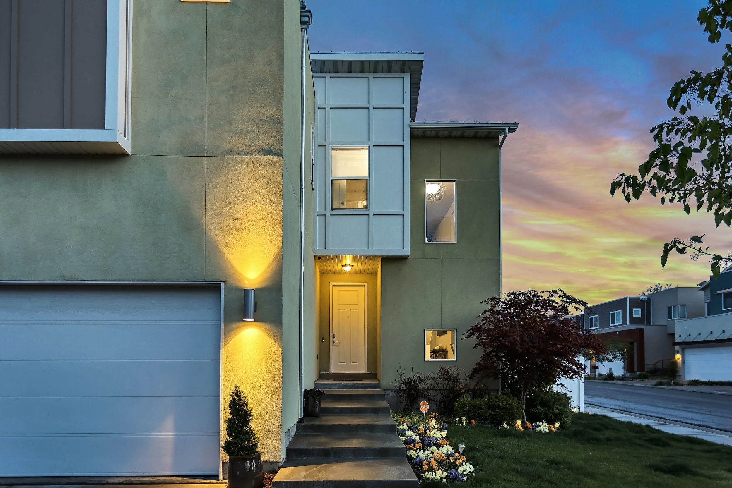 Modern multi-story house with a yellow exterior, front steps leading to a white door, illuminated by exterior lighting, with a garden of flowers and bushes, and neighboring houses visible in the background during dusk.