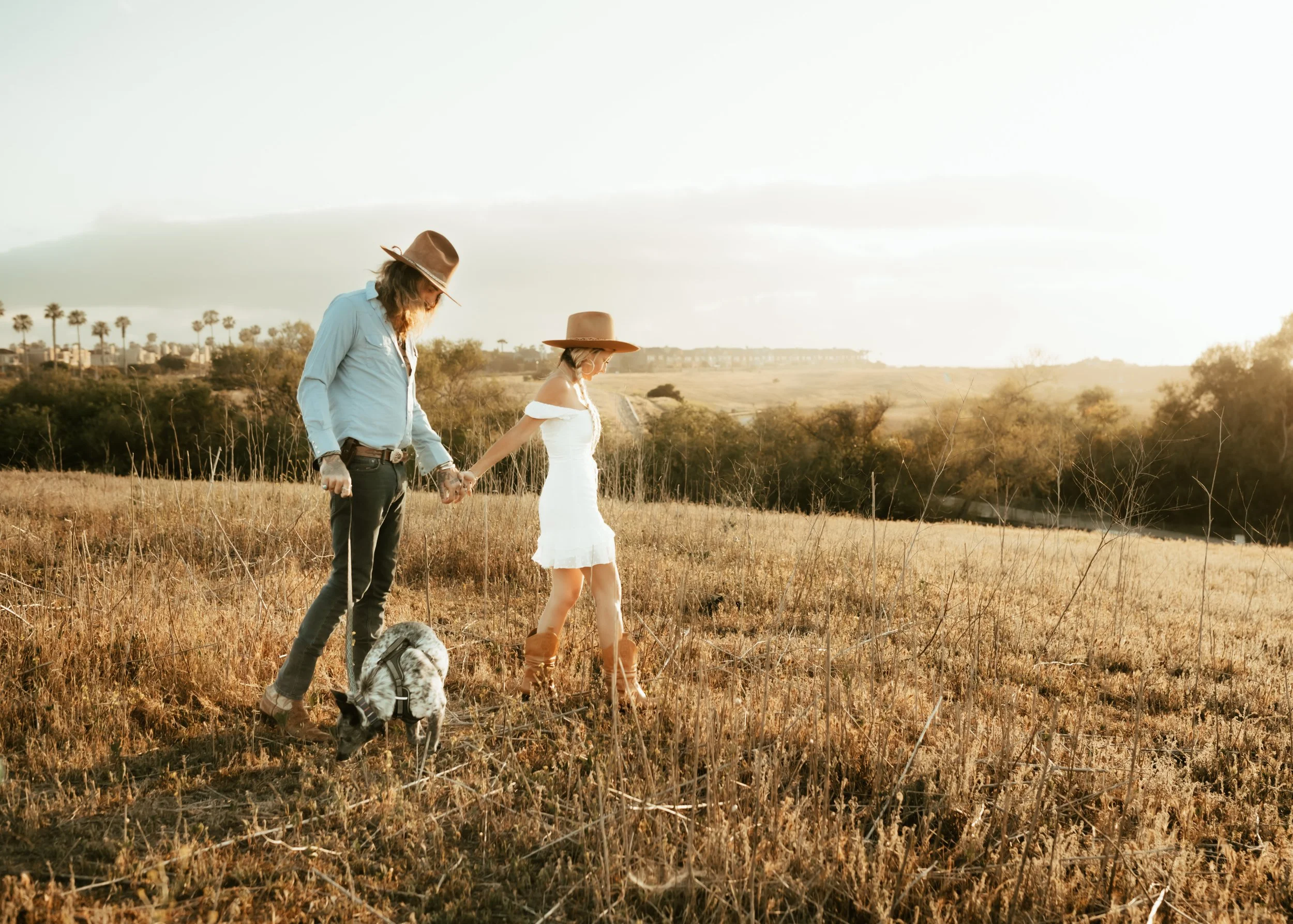 A couple holding hands while walking through a field with their dog during sunset, with palm trees and a distant city skyline in the background.