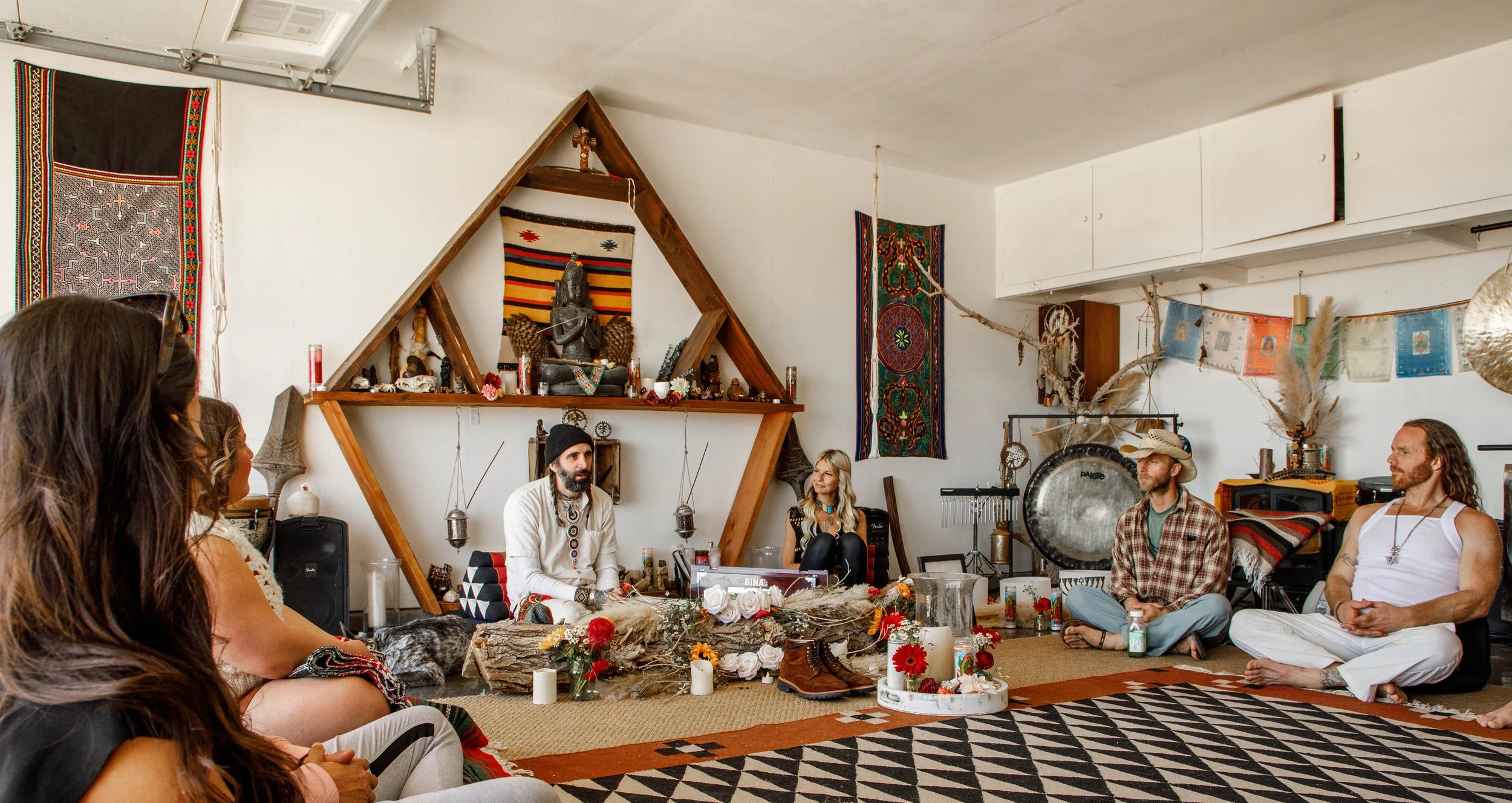 A group of people sitting on the floor in a circle during a spiritual or ceremonial gathering in a decorated room with tapestries, candles, and a central altar with a statue and offerings.