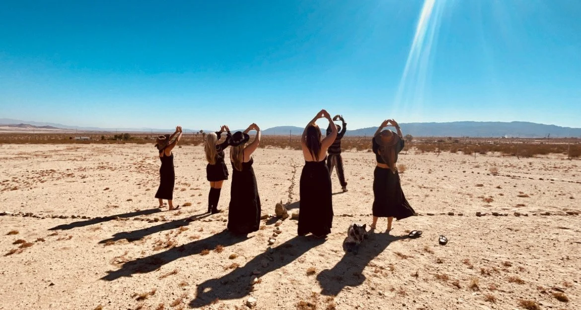 Group of six women in black dresses and wide-brimmed hats standing in a desert, facing away, with mountains in the background and shadows cast on the ground.