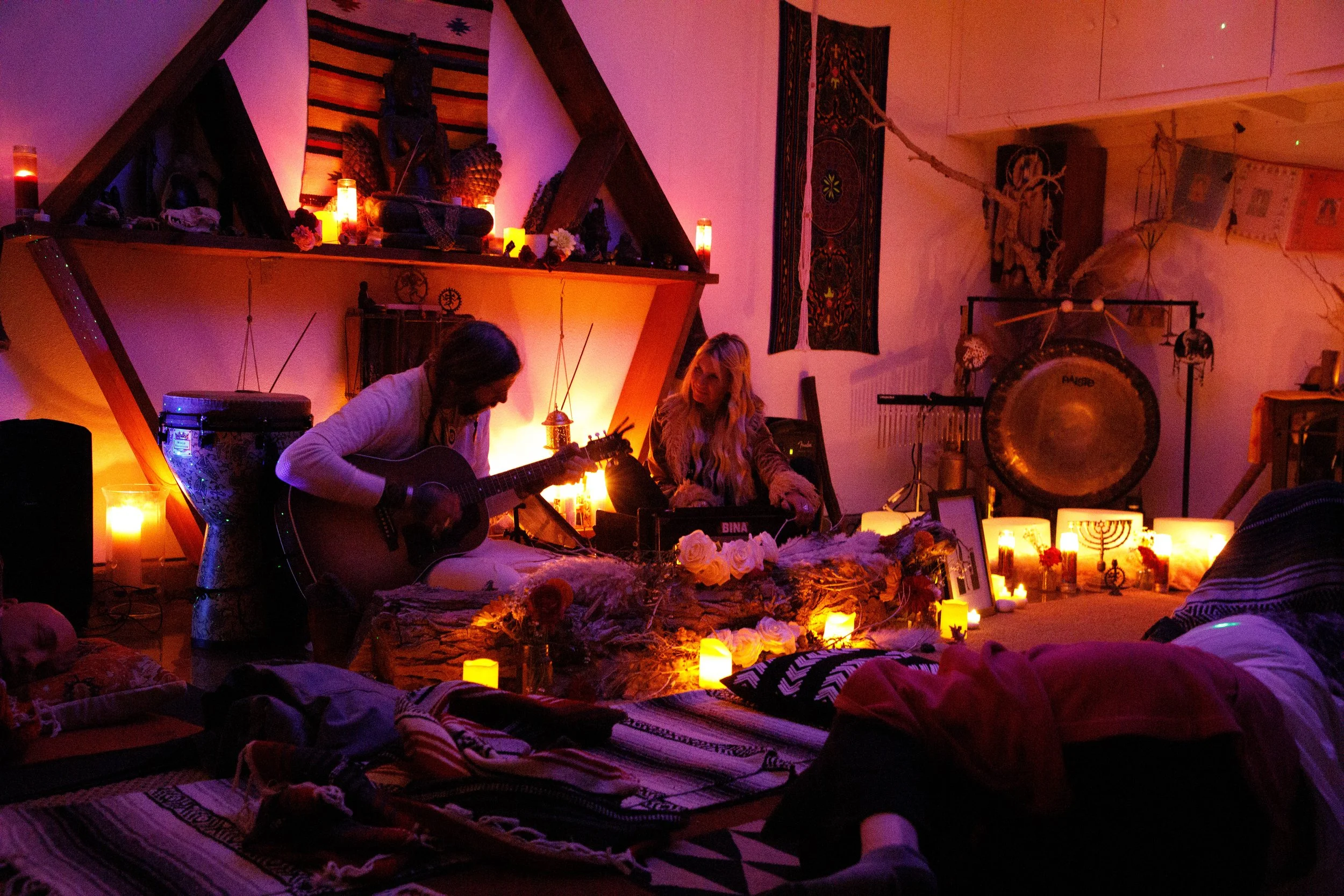 Two women perform in a dimly lit room filled with candles, flowers, and bohemian decor. One is playing a guitar while the other is using a sound mixer, surrounded by spiritual and artistic items.