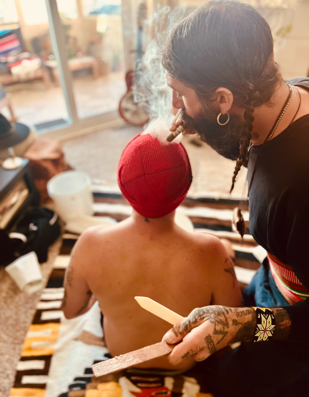 A person with tattoos and a red beanie smoking a cigar while sitting on a striped blanket, as another person with braided hair and tattoos uses a tattoo machine on their back in a cozy indoor setting.