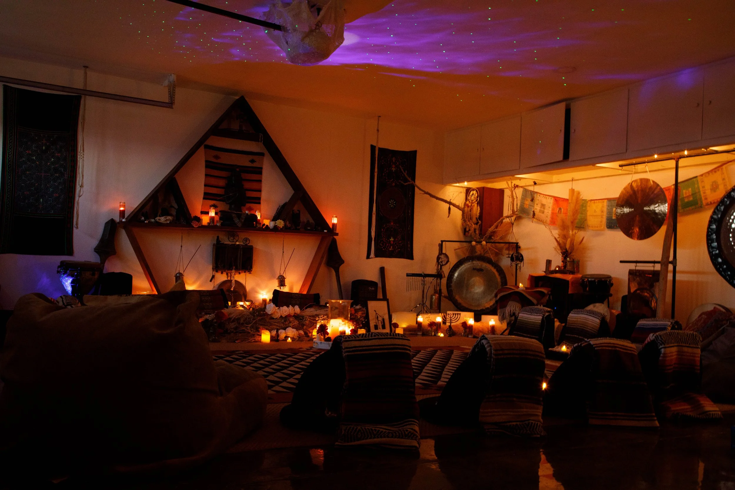 A dimly lit room decorated with candles and musical instruments, including gongs and drums. The ceiling has a projection of colorful lights. There is a triangular altar with candles and statues on a shelf, and various textiles and tapestries adorn th