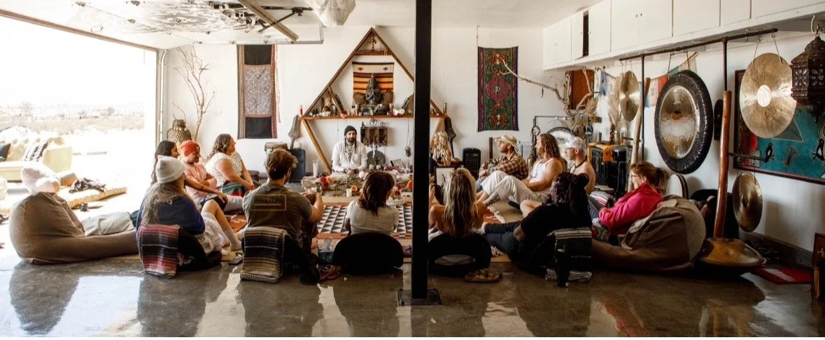 People sitting on the floor in a circle participating in a meditation or yoga session in a decorated indoor space with gongs and artwork.
