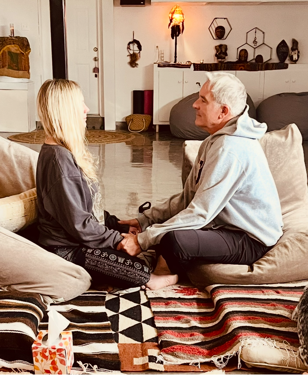 A young woman and an older man are sitting on cushions on the floor facing each other, holding hands, and engaging in a close, intimate conversation in a cozy living room with warm lighting and decorative items on the wall.
