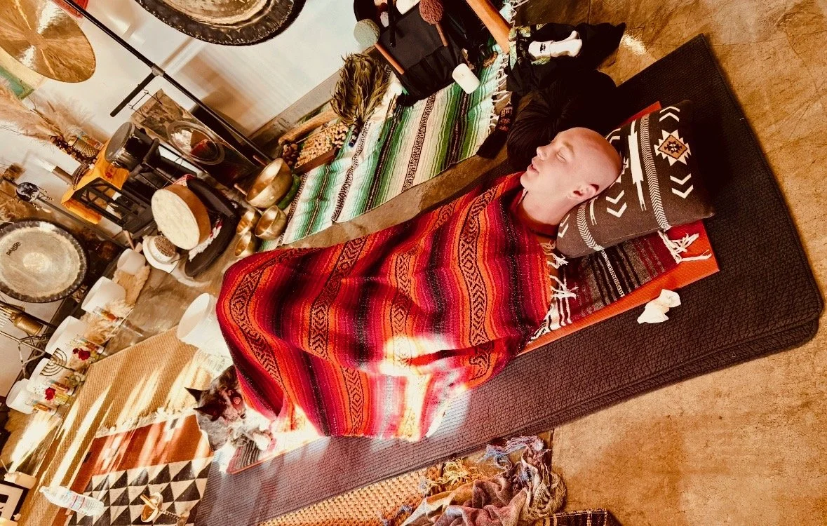 A person lying peacefully on a mat with eyes closed, covered with a colorful blanket, resting on a pillow, in a cozy indoor setting with music equipment and decorative items around.