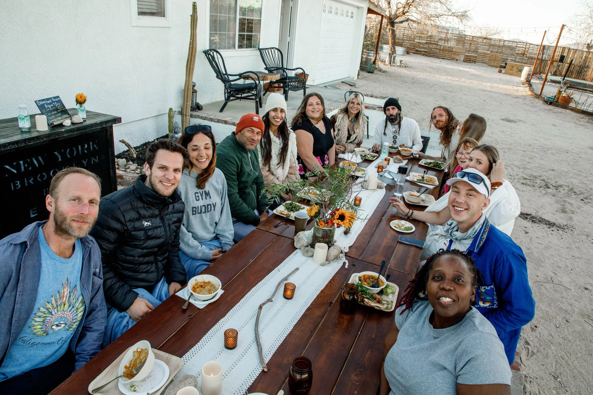 Group of people gathered around a long outdoor dining table, enjoying a meal. The table is decorated with a floral centerpiece and candles, set on a rustic tablecloth. The setting appears to be in a backyard or patio area with a sandy ground, a house