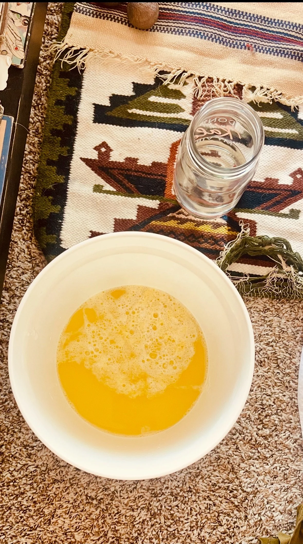 A glass filled with water and a white bowl containing a yellow liquid, possibly egg mixture, on a patterned fabric tablecloth.