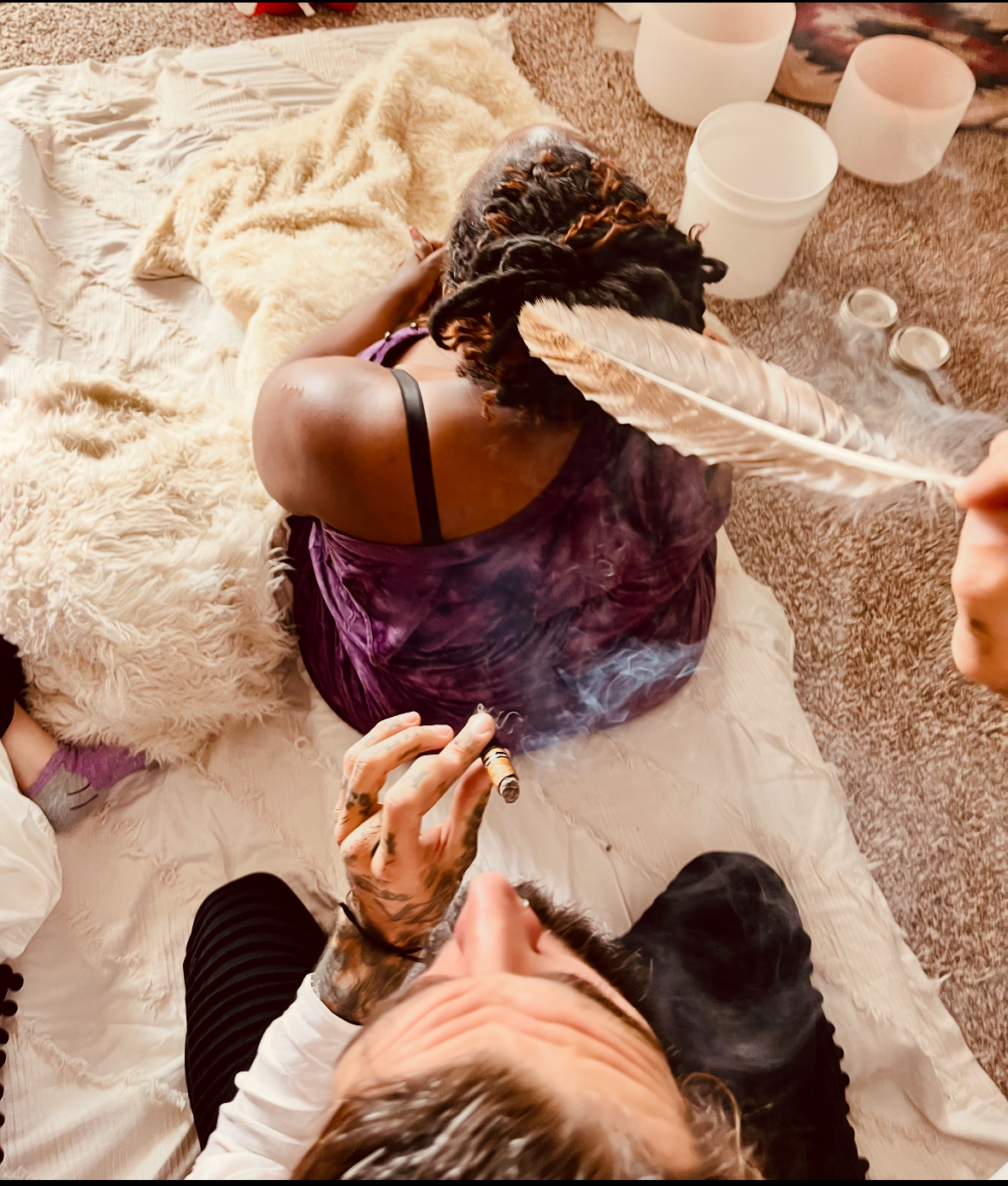 Person lying on a bed with furry blankets, holding a black dog, while another person leans over with a feather in hand. There are large white candles on the floor and a lit cigar in the person's hand.