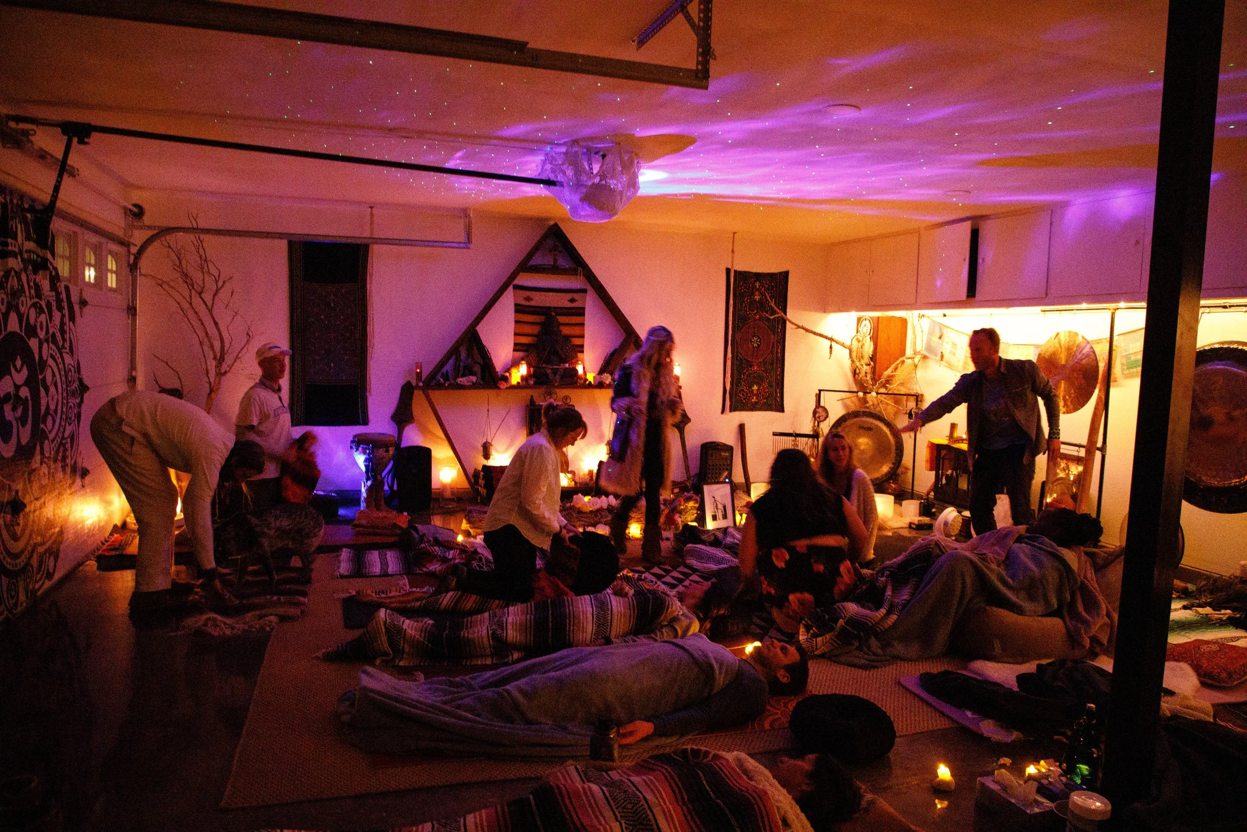 People participating in a spiritual or meditation gathering in a cozy, dimly lit room decorated with tapestries, candles, and spiritual artifacts. Some are sitting or lying on mats, while others are standing or engaging in conversation. The room is i