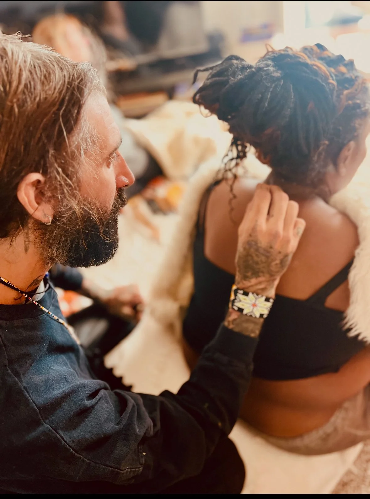 A man with long hair and a beard is touching a woman's shoulder in a close-up shot. The woman has dark, curly hair and is wearing a black top with a white fur coat draped over her. The background shows other people seated in a blurred setting.