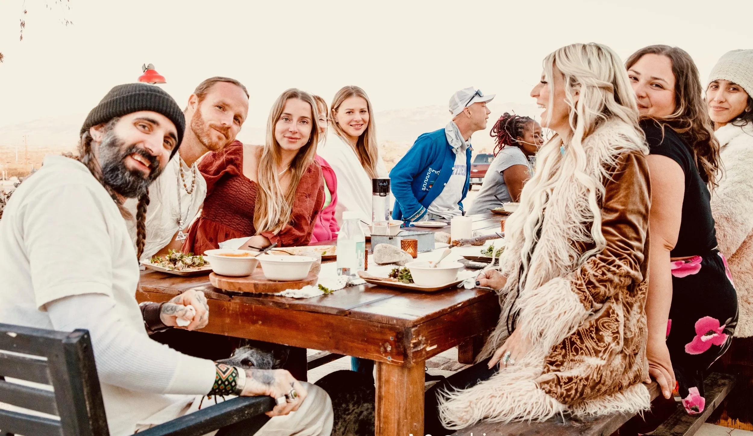 A group of people gathered outdoors around a wooden table, sharing a meal and smiling.