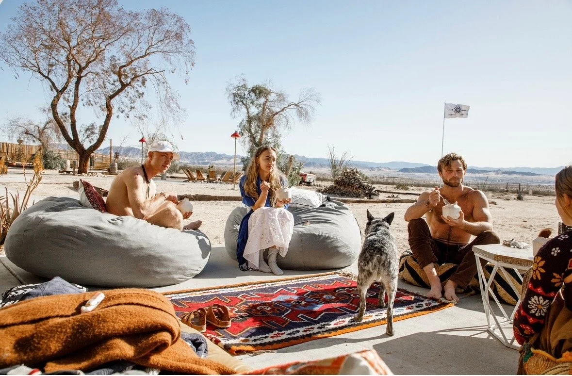 Group of four people and a dog relaxing outdoors on a sunny day in a desert setting, with some trees and mountains in the background. The people are sitting on outdoor cushions and a rug, eating from bowls.