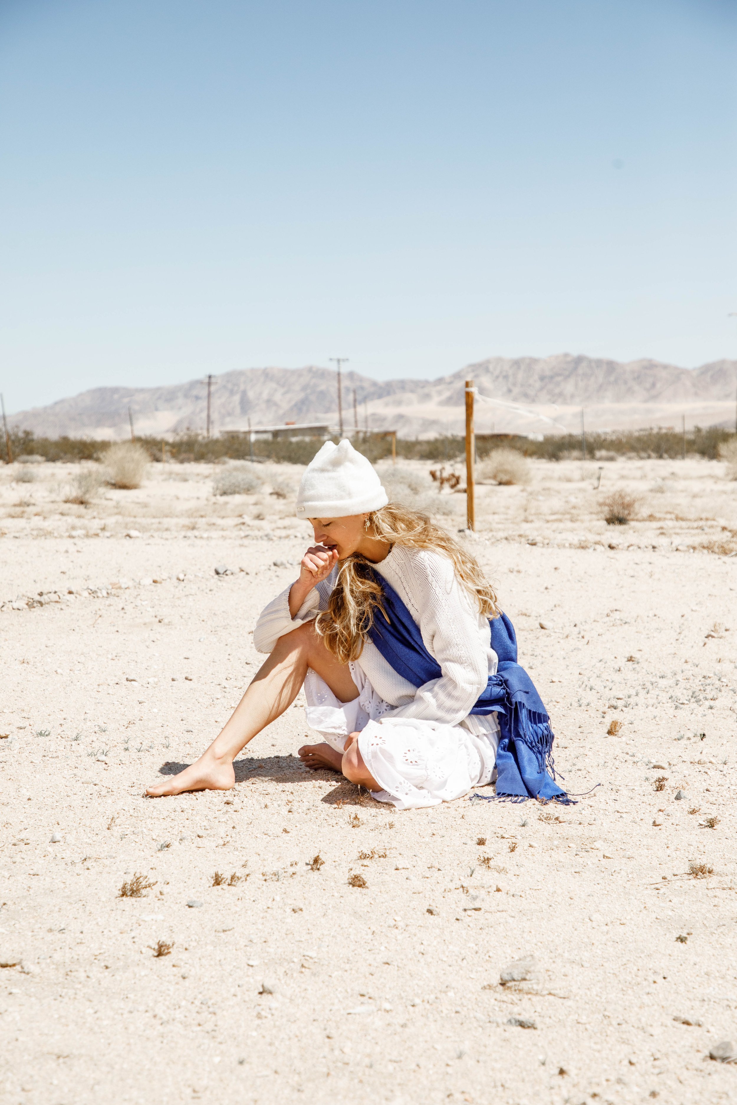A woman in a white hat, white dress, and blue shawl sitting on sandy desert ground, with mountains in the background under clear blue sky.