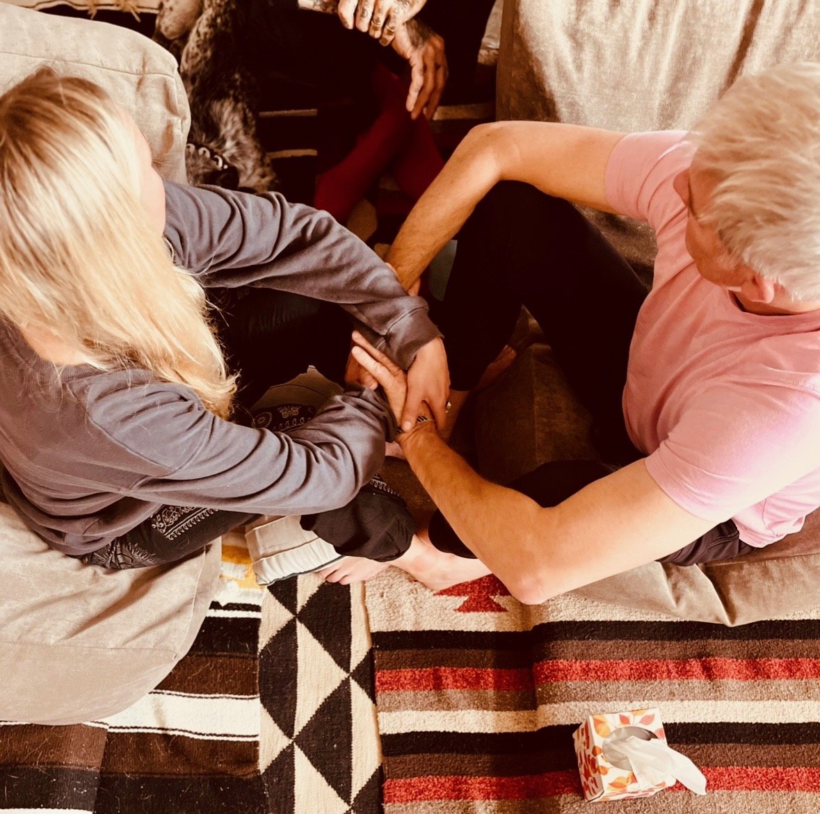 Two people, a man with blond hair in a pink shirt and a woman with blonde hair in a gray jacket, are sitting on a couch and engaging in a playful arm wrestling match with each other. A tissue box is on the floor nearby.