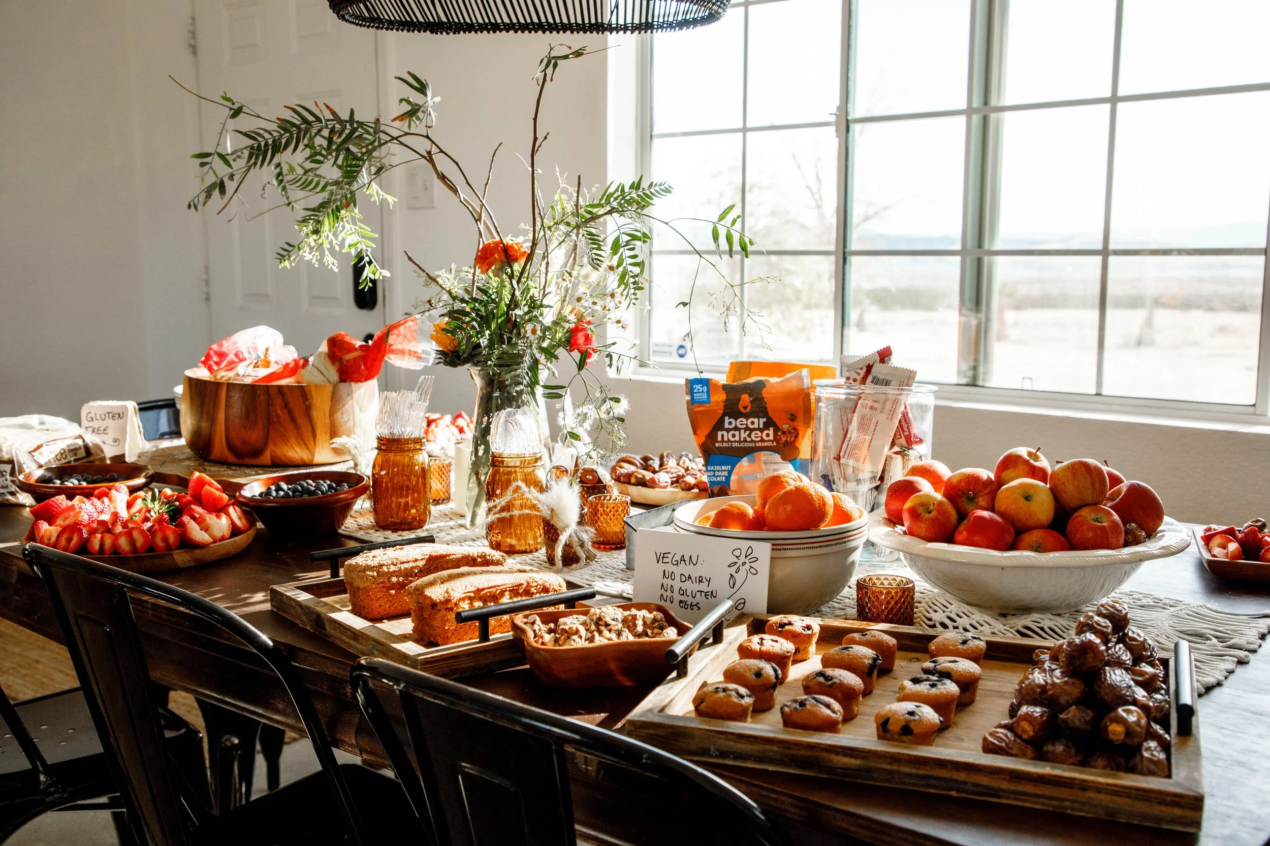 A table filled with various fruits, baked goods, and snacks, with a vase of flowers in the center, near a window letting in natural light.