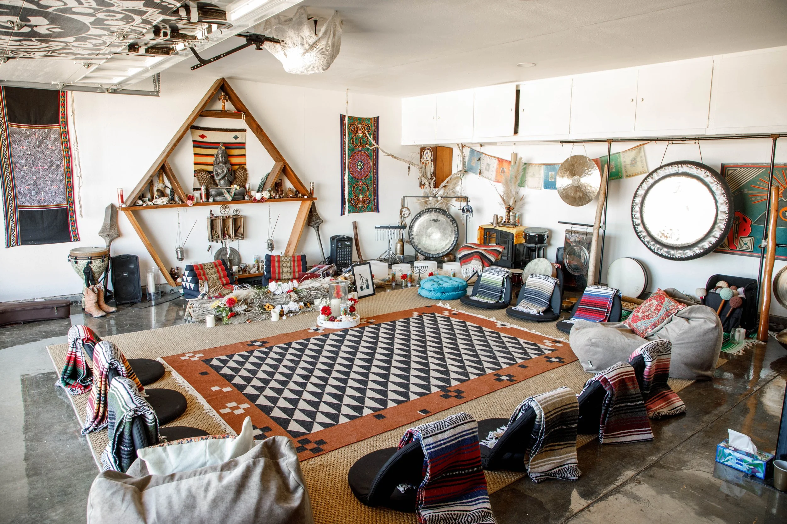 A decorated indoor space with musical instruments, shelves, and a patterned rug. Chairs and cushions are arranged around the rug, with various decorative items and fabrics on the walls and furniture. Gongs, cymbals, and other percussion instruments a