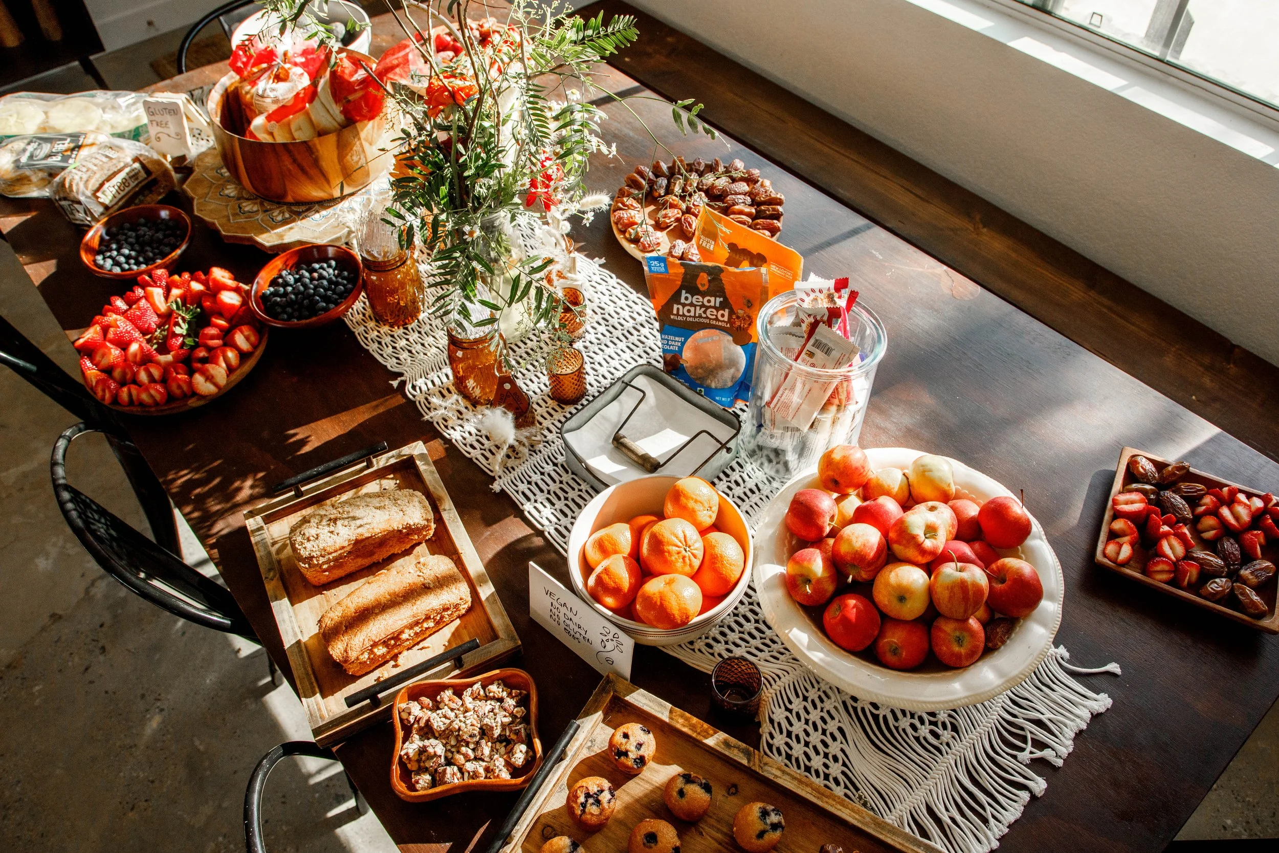 A dining table with various fresh fruits such as strawberries, blueberries, apples, and oranges, along with baked goods, snacks, and a floral centerpiece, set near a window with sunlight streaming in.