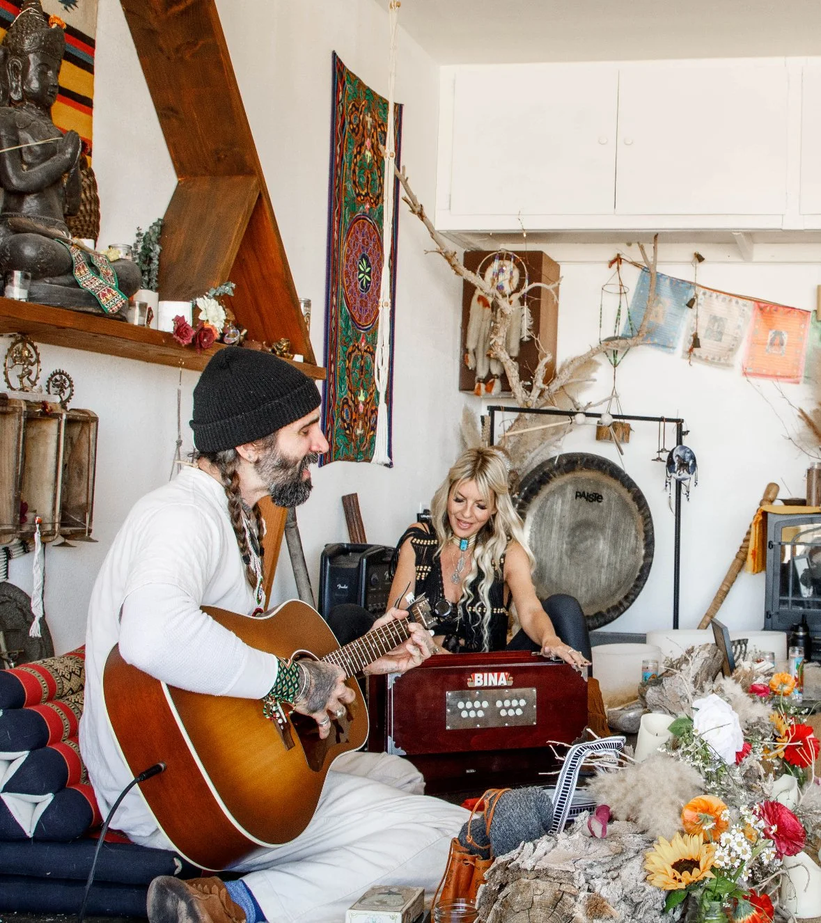 Two people playing music indoors, a man with a guitar and a woman with a harmonium, surrounded by eclectic decor and flowers.