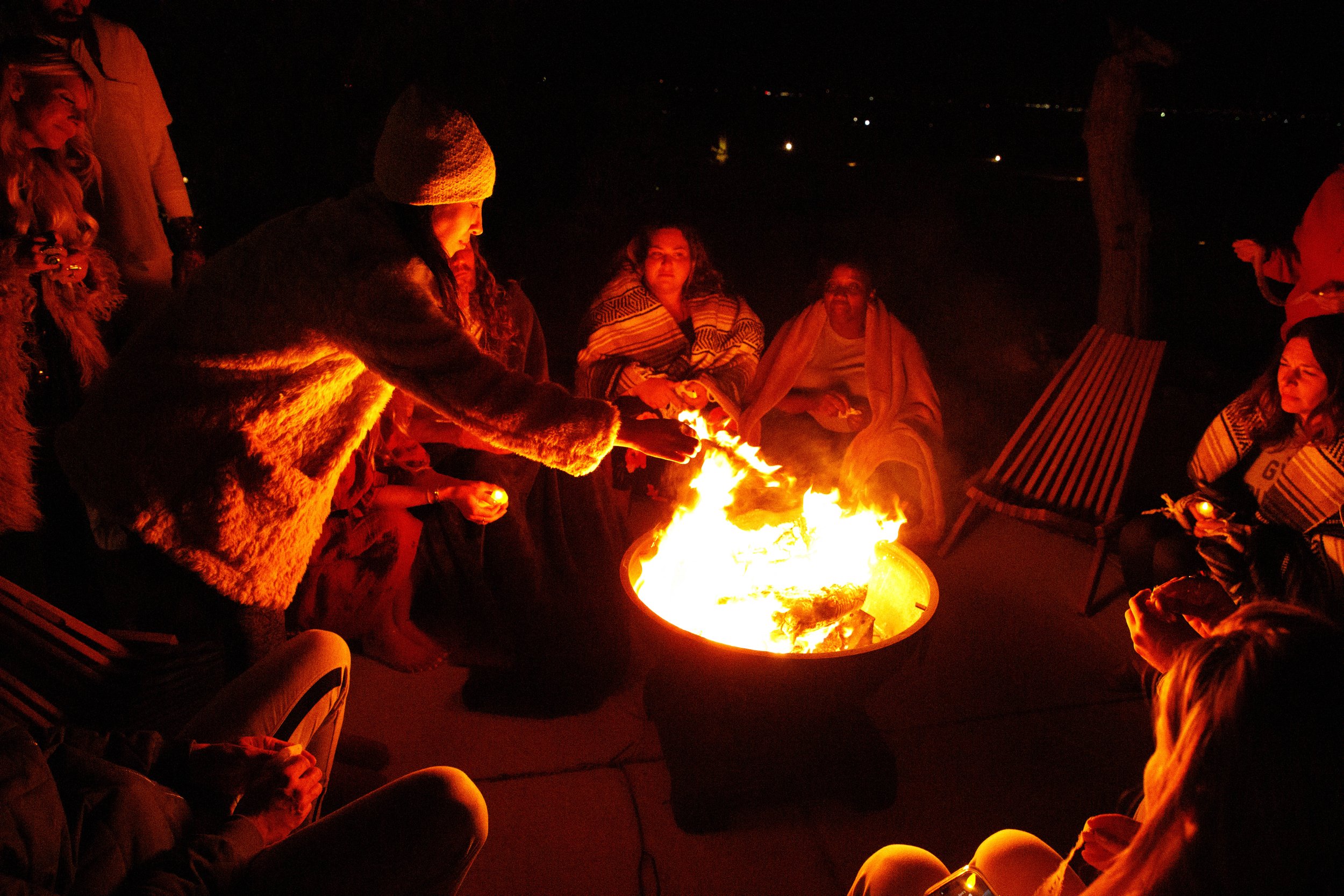 People gathered around a fire pit outdoors at night, some sitting on chairs and some standing, with city lights in the background.