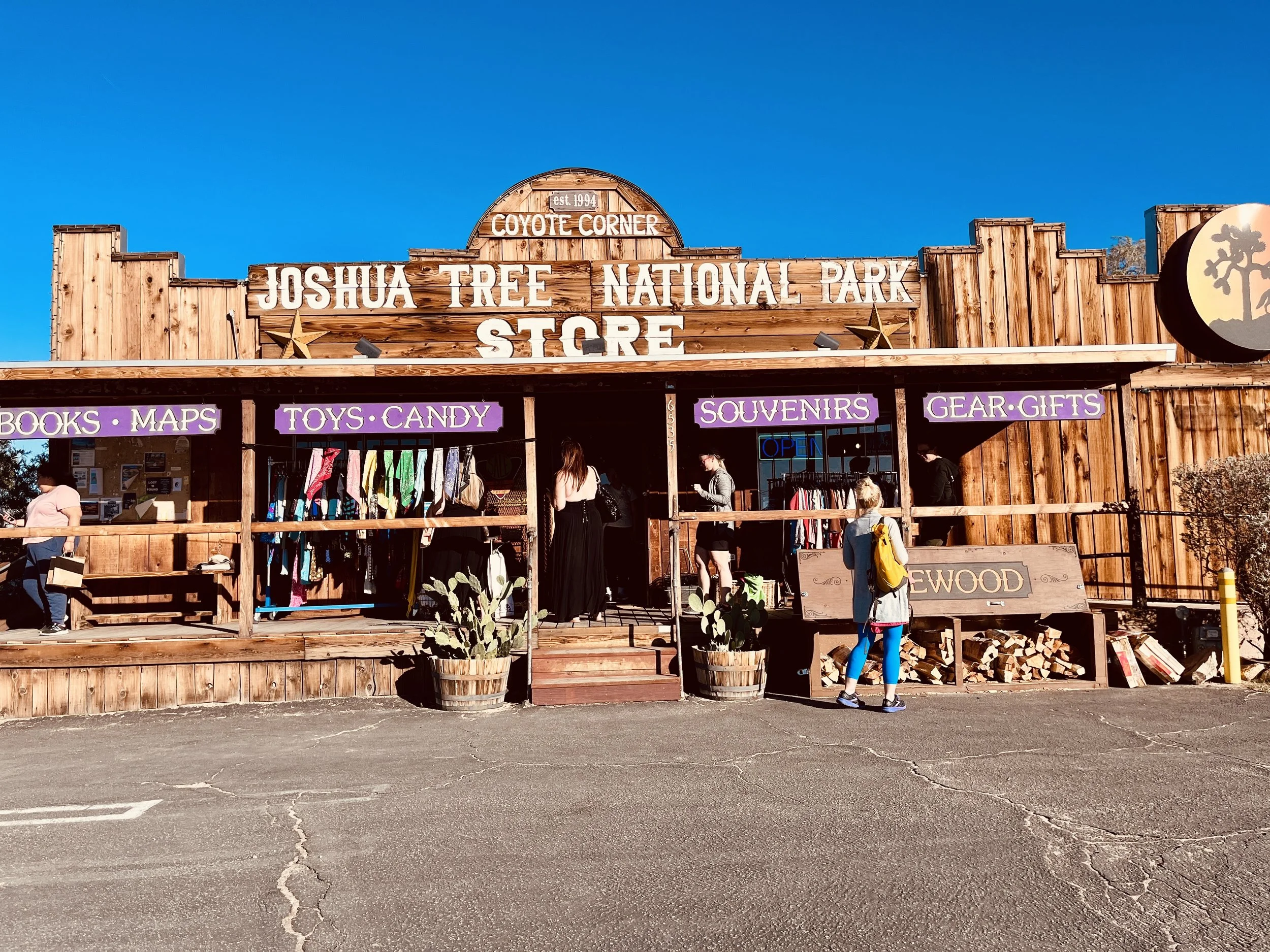Wooden store building labeled "Joshua Tree National Park Store" with purple signs for books, maps, toys, candy, souvenirs, gear, and gifts, and people shopping outside on a sunny day.