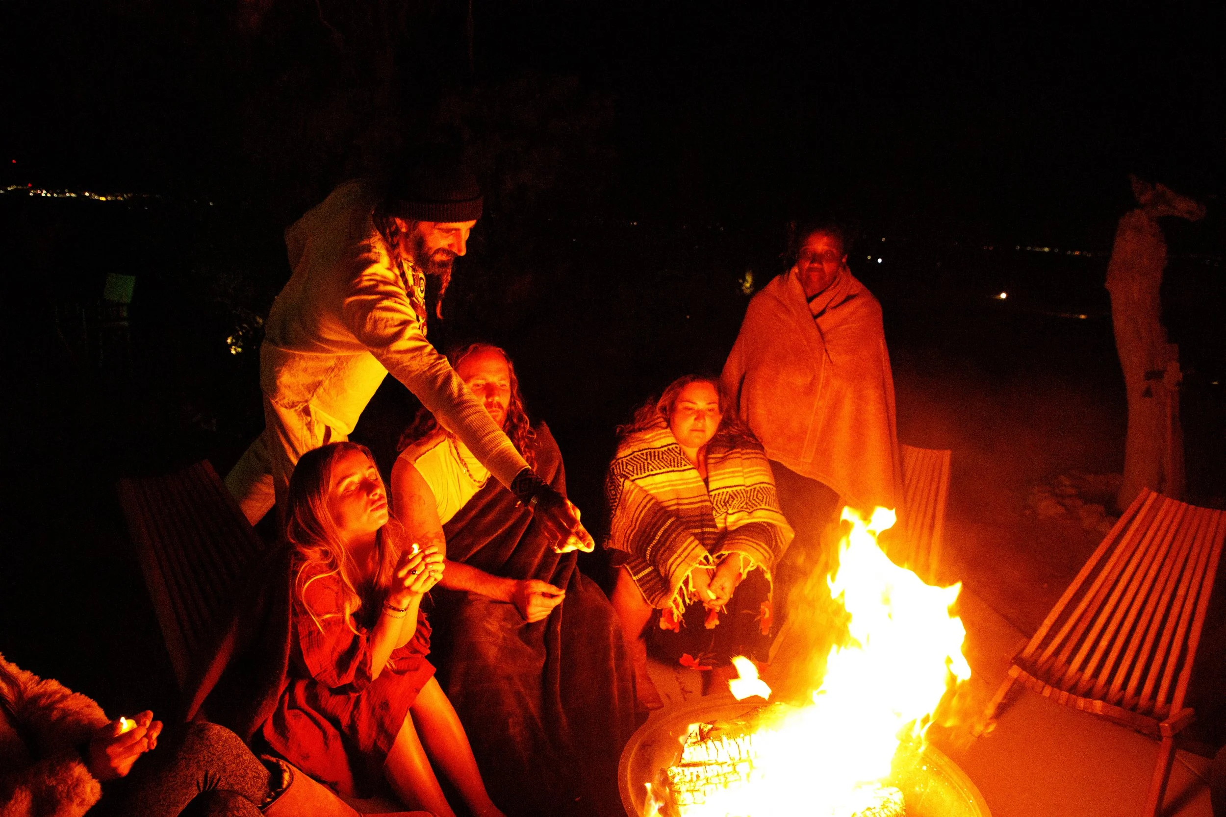 Group of six people sitting and standing around a campfire at night outdoors, with some seated on chairs and wrapped in blankets, enjoying the fire.