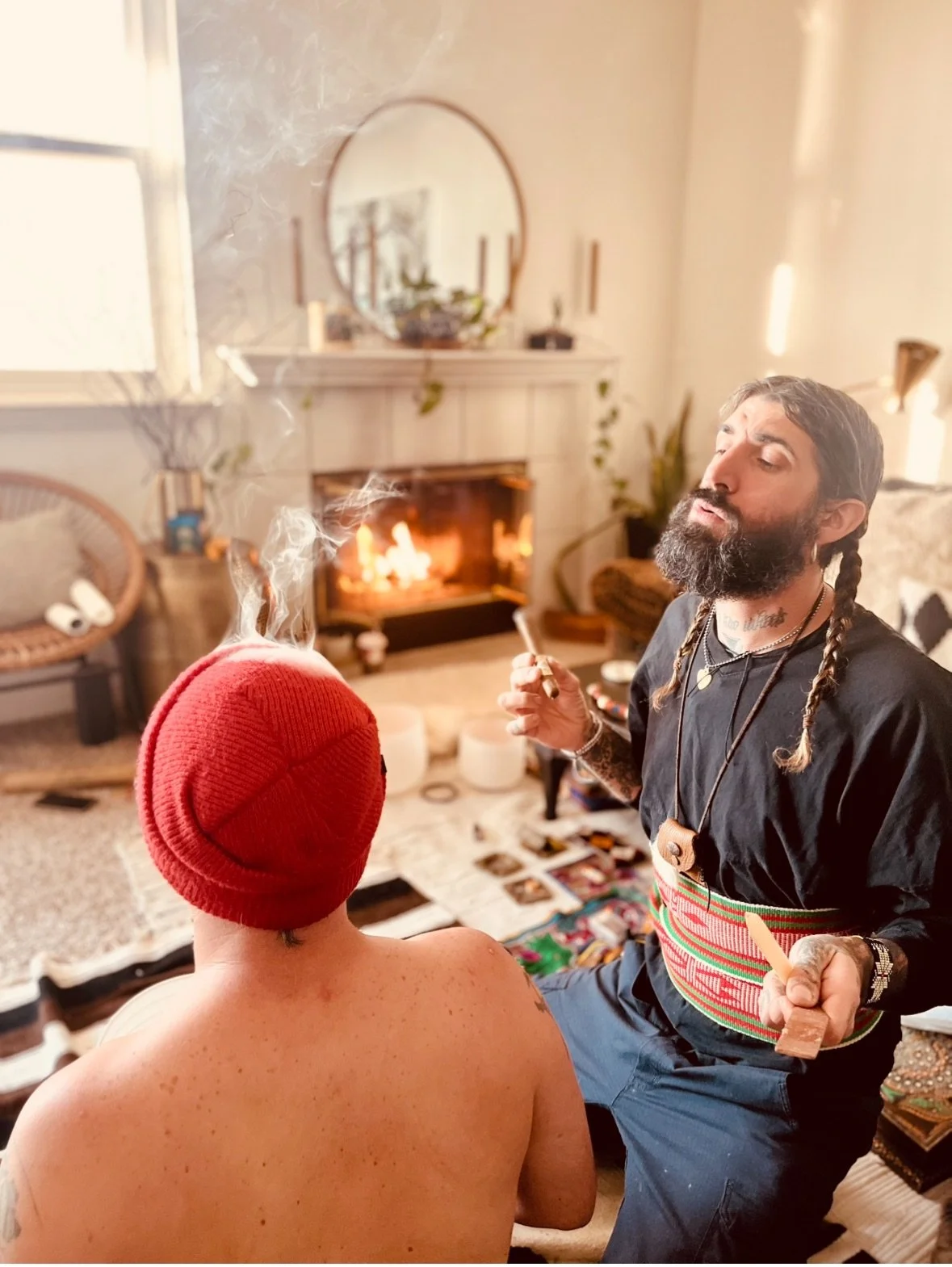 A man with a beard and braids sitting shirtless in a living room, wearing a red beanie, talking to a woman with a red beanie and no shirt, smoking a cigarette, with a fireplace in the background.