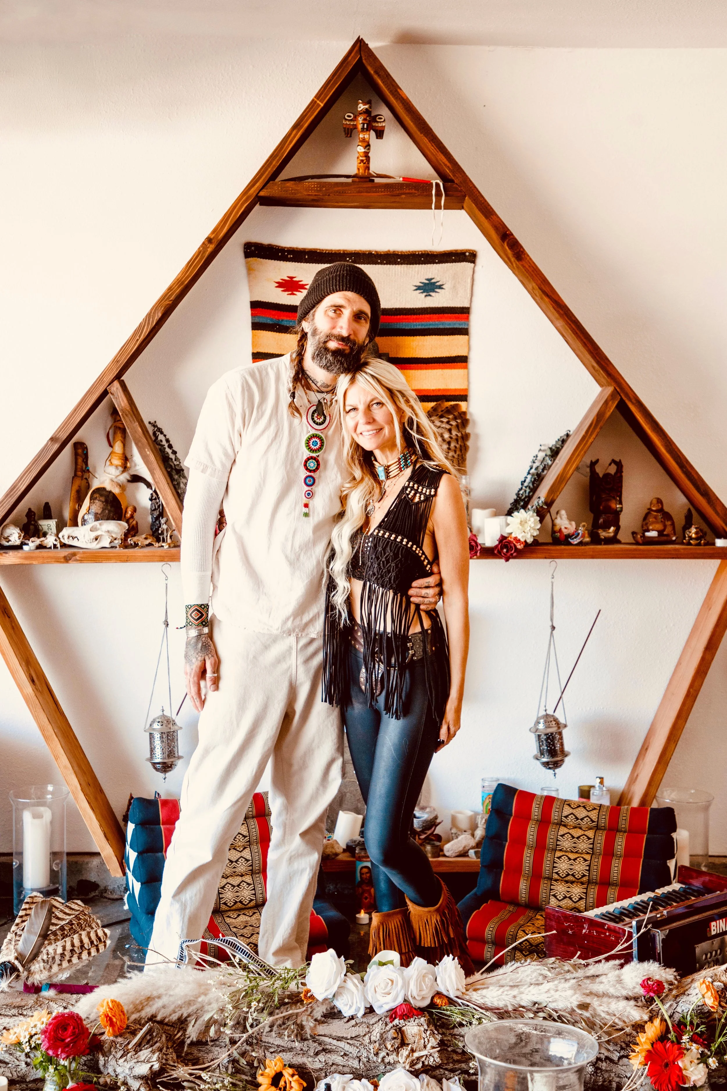 A man and woman standing together inside a geometric wooden shelf decorated with tribal and spiritual items, surrounded by flowers, candles, and patterned cushions.