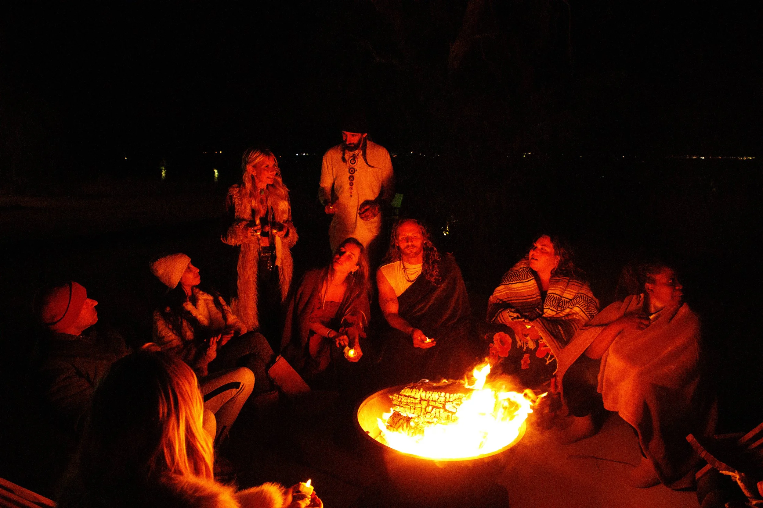 Group of friends sitting around a campfire at night, some holding drinks, with a dark sky and distant lights in the background.