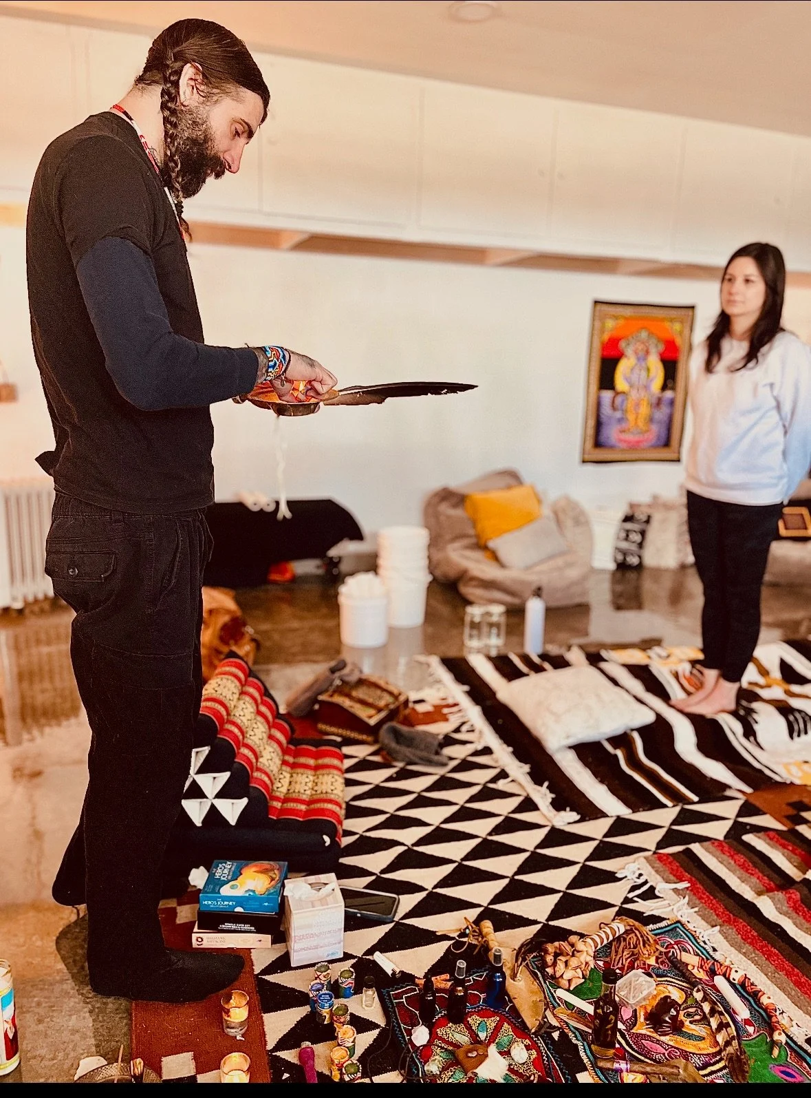 A man with long hair and beard wearing a black shirt with colorful bracelets, holding a feather and a bowl, standing on a patterned rug with candles and bottles. A woman in a white hoodie and black pants stands in the background in a room with a colorful portrait on the wall, cushions, and various objects on the floor.