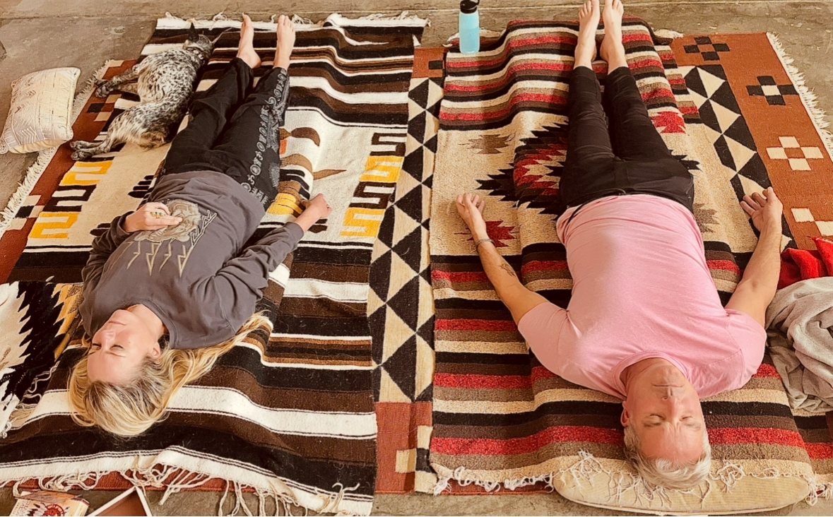 Two people lying on colorful Southwestern-patterned blankets on the floor with their eyes closed, next to a sleeping cat and a water bottle.