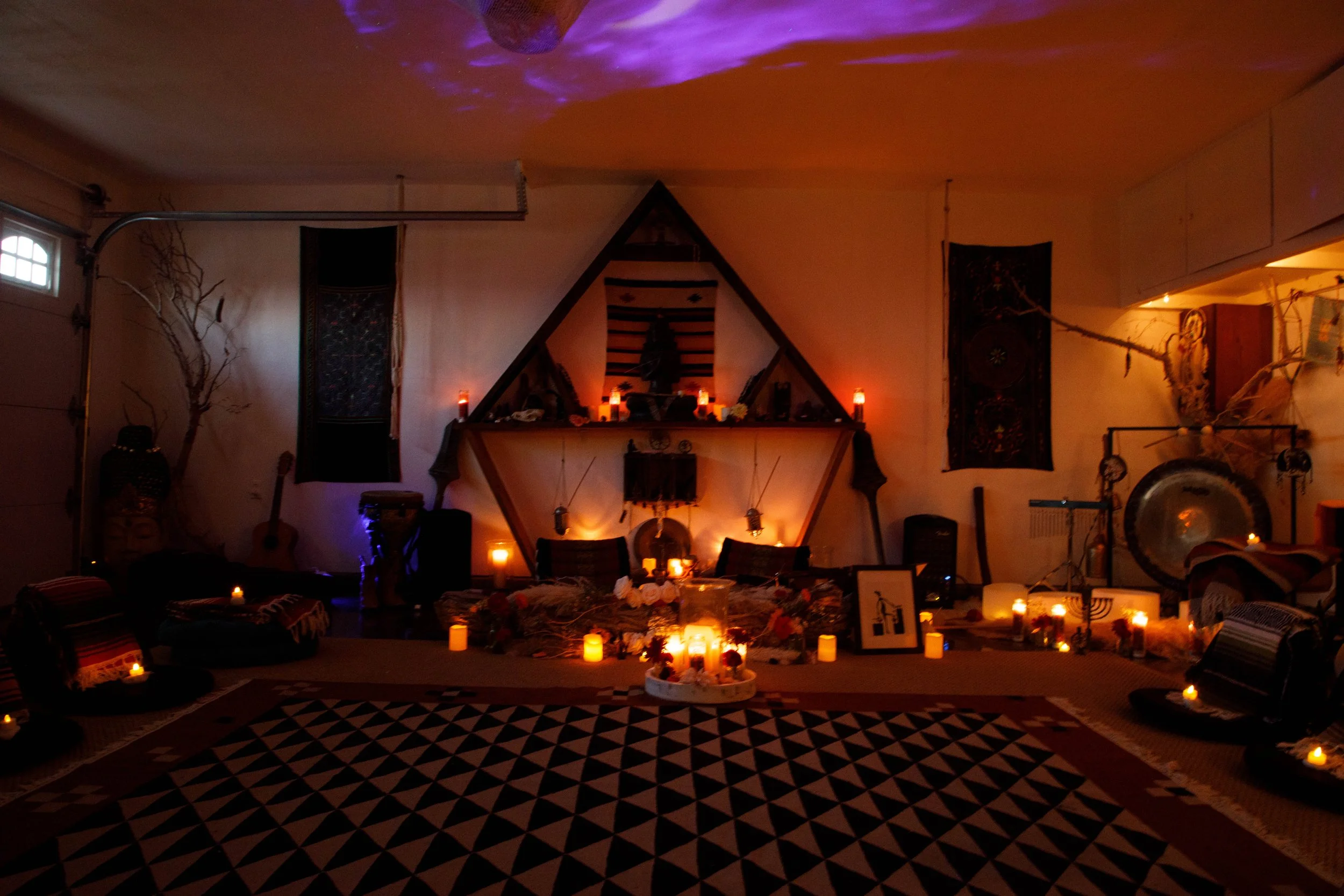 A dimly lit room decorated with numerous candles, a large rug with geometric pattern, musical instruments, and mystical or spiritual items, creating an atmosphere of an altar or meditation space.