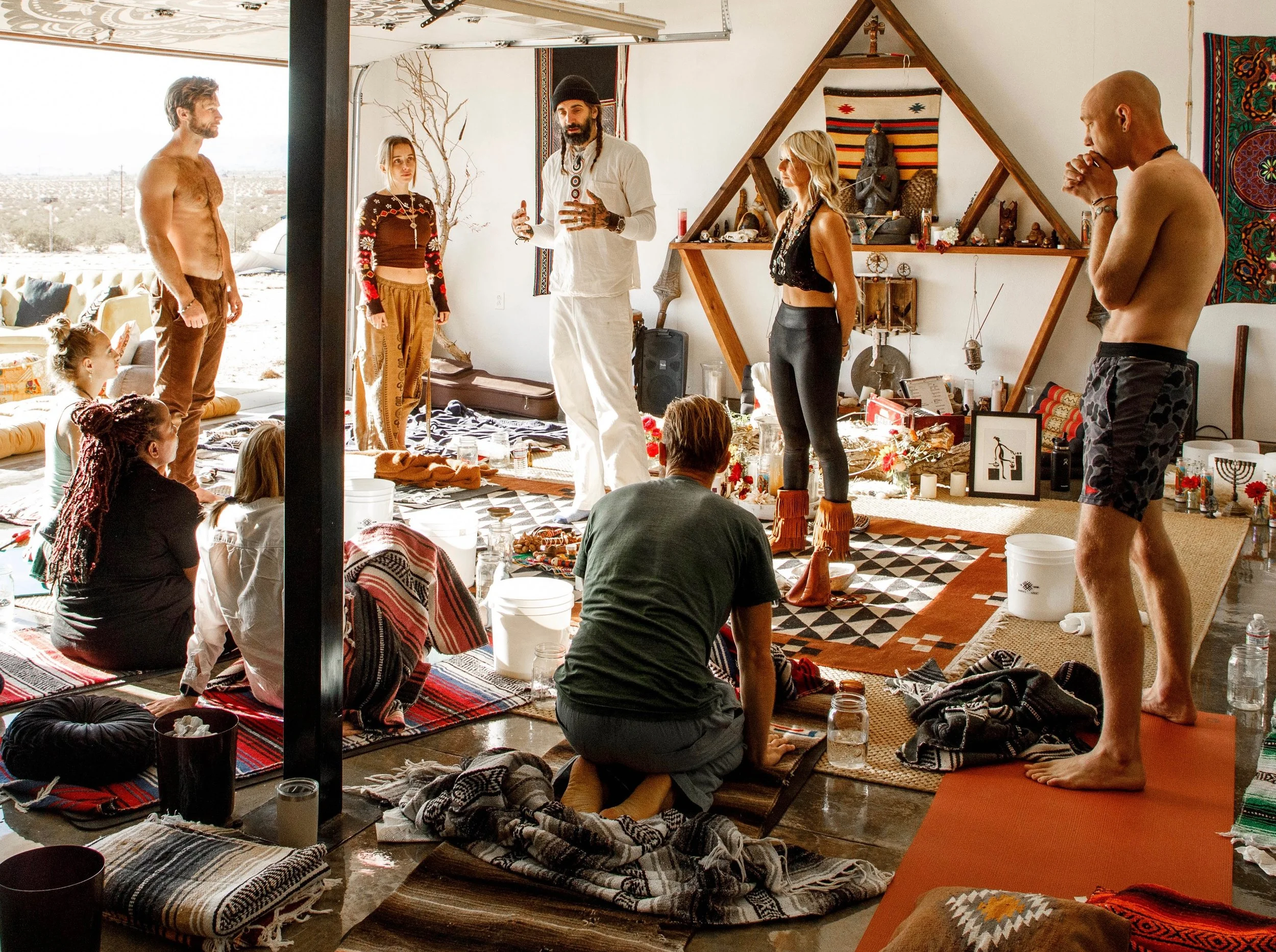 Group of people participating in a meditation or spiritual gathering in a bright, decorated room with various spiritual artifacts, woven textiles, and a large window showing a desert landscape outside.
