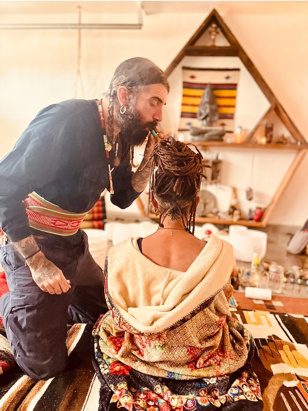 A man with tattoos, earrings, and long hair and beard being blessed by a woman with dreadlocks and tribal clothing in a cozy, decorated room.