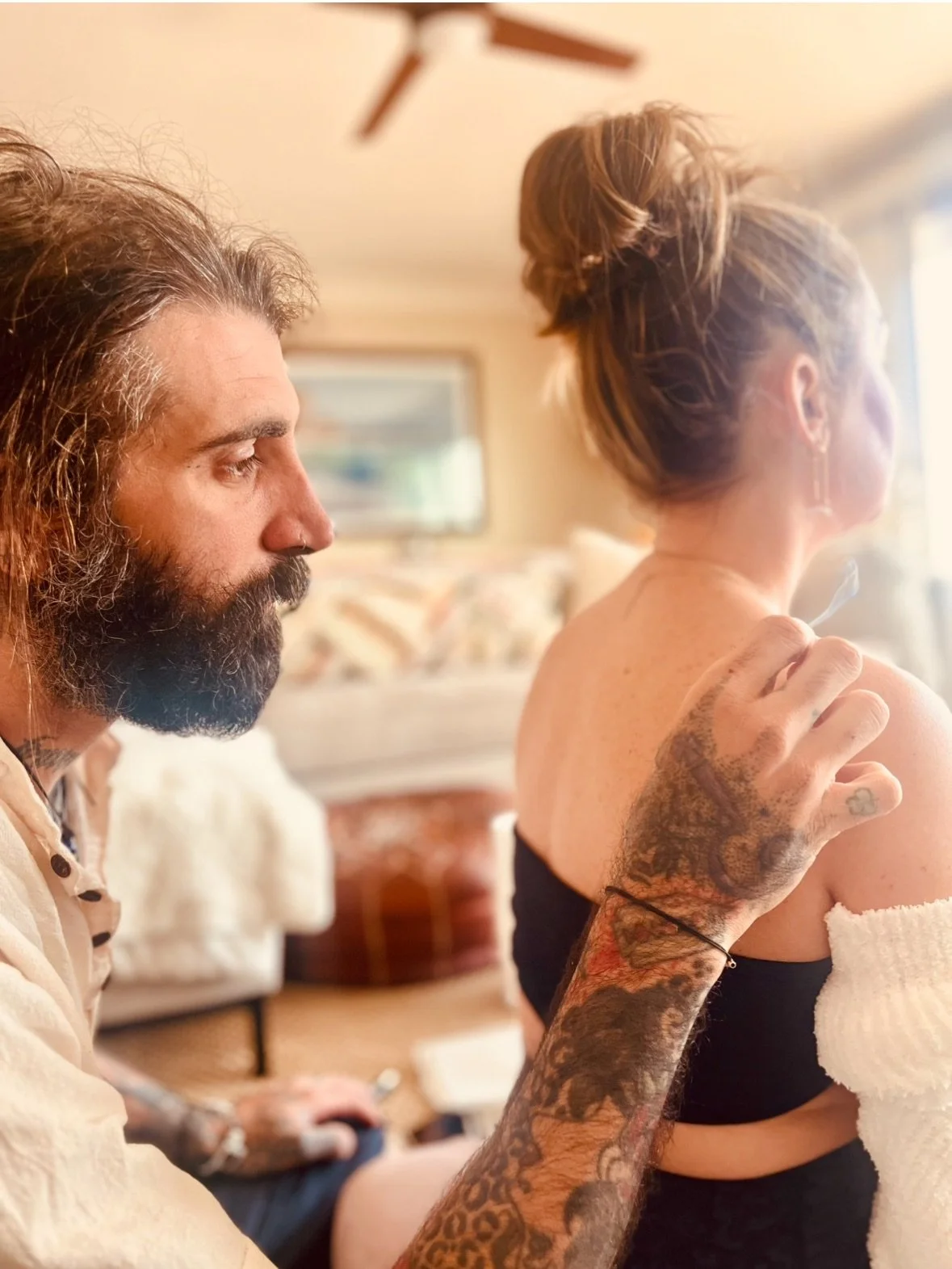 Tattooed man with long hair and beard, gently touching a woman's shoulder in a cozy, well-lit room.