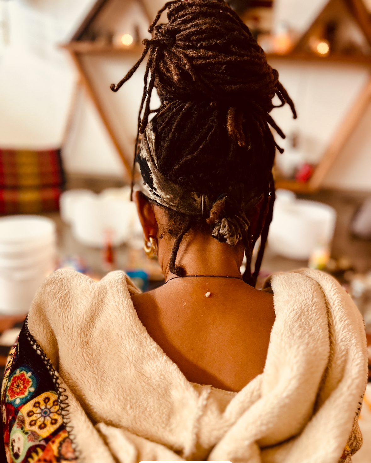 Rear view of a woman with styled dreadlocks in a bun, wearing earrings and a fleece-lined garment, with a blurred shelf and colorful items in the background.