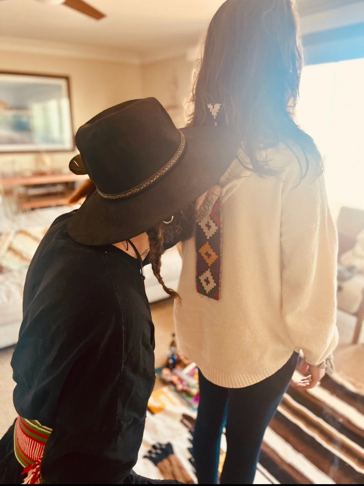 Person with long dark hair standing next to another person, who is wearing a large floppy hat with a gold chain band, in a cozy room with a patterned rug, a table, and a picture on the wall.